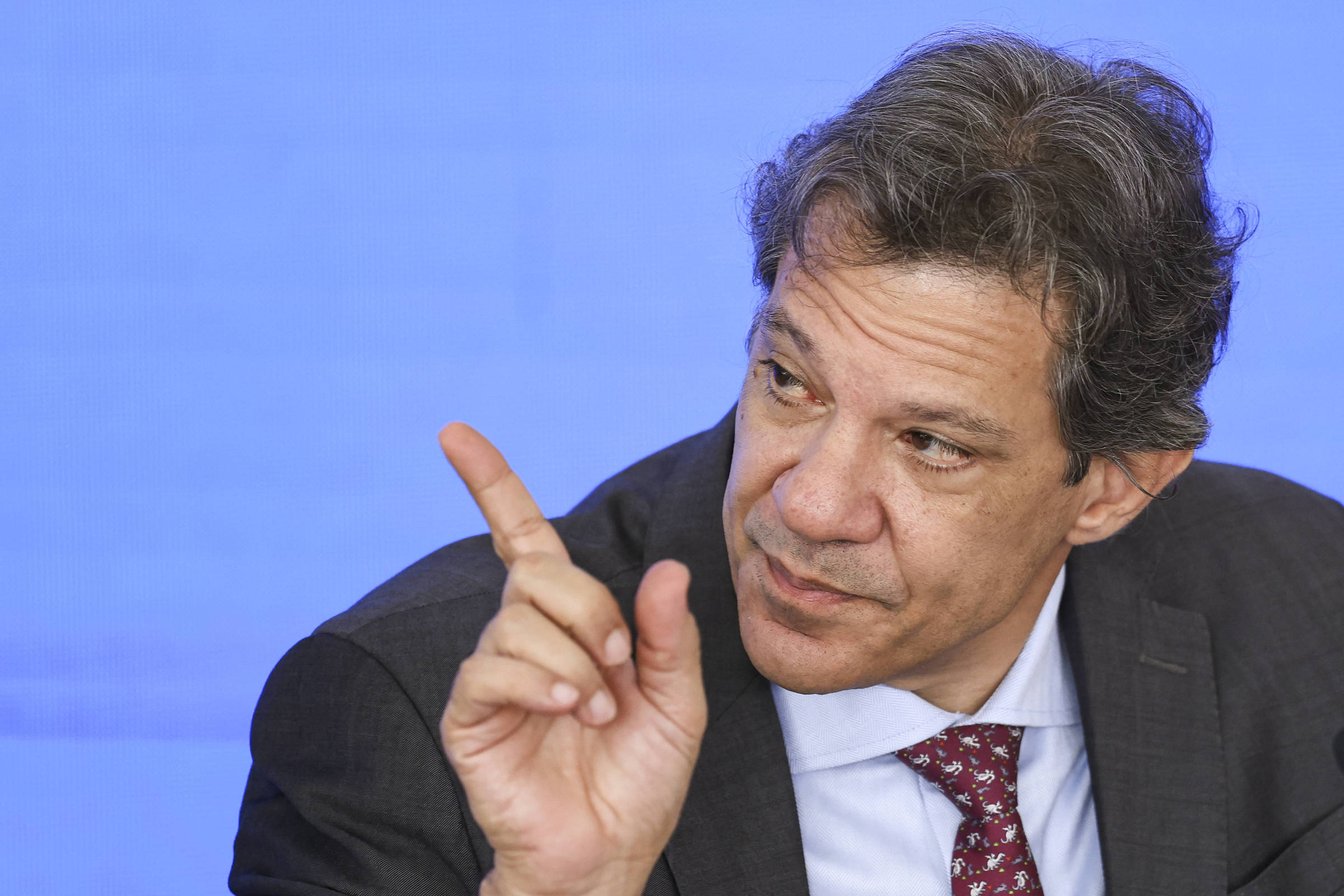Um homem com cabelo castanho e levemente ondulado está sentado, vestindo um terno escuro e uma gravata vermelha com padrões. Ele está gesticulando com a mão direita levantada, enquanto olha para a frente. O fundo é azul claro.