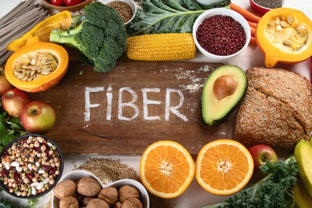 Alimentos com fibras como frutas e legumes em cima de mesa de madeira e ao meio está escrito fiber de giz branco