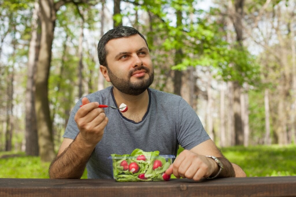 Homem sentado em frente a uma mesa de madeira e ao ar livre comendo salada com rabanete