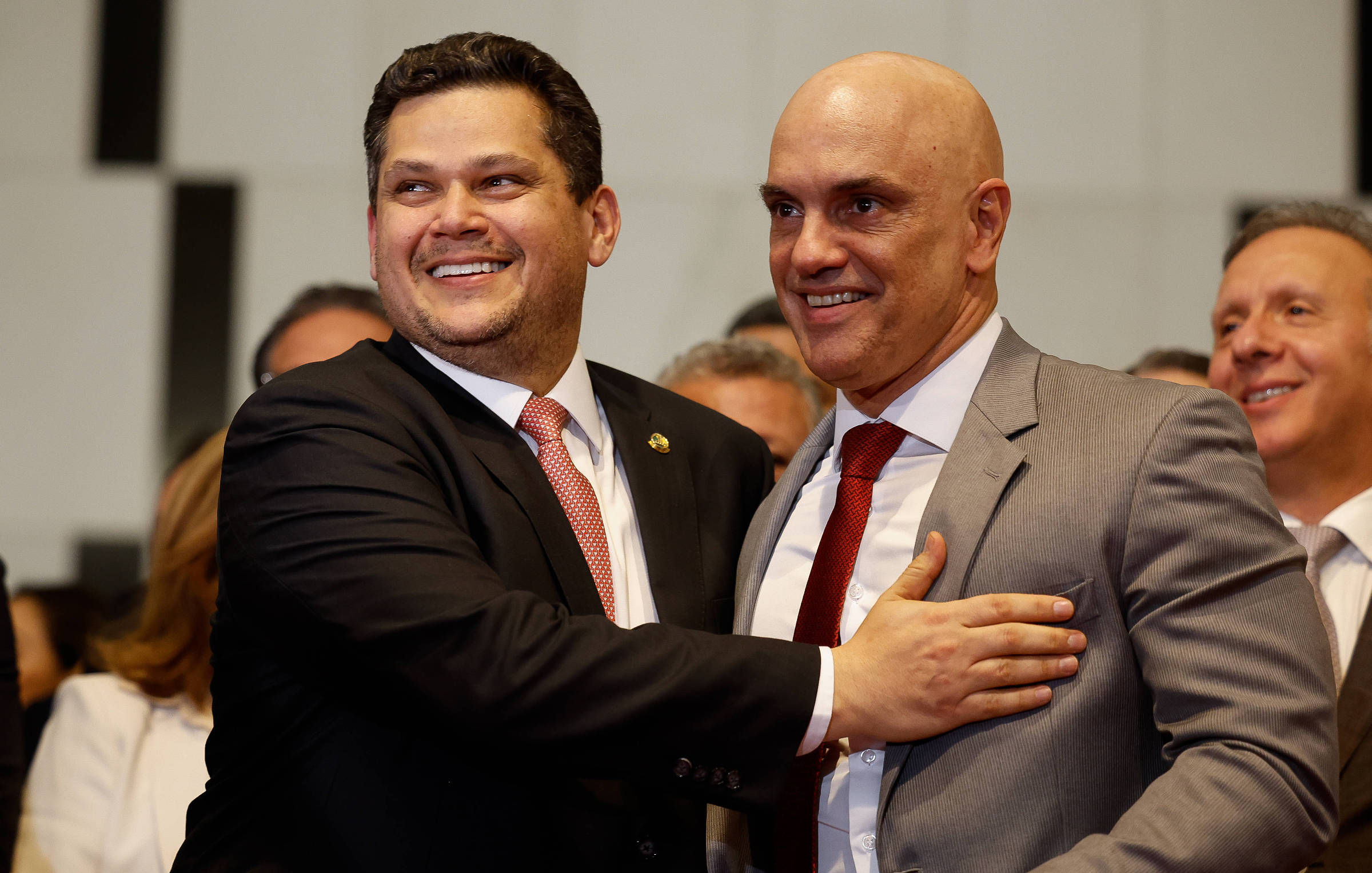 A imagem mostra dois homens sorrindo e se cumprimentando em um evento. O homem à esquerda está vestindo um terno escuro com uma gravata vermelha e uma camisa branca, enquanto o homem à direita usa um terno cinza claro com uma camisa branca e uma gravata vermelha. Ao fundo, há outras pessoas sorrindo e observando a cena.