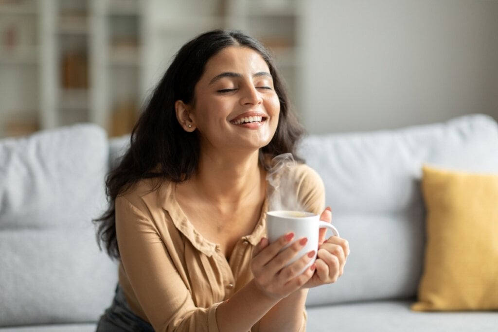Mulher sorrindo com os olhos fechados, sentada em um sofá, e segurando uma xícara de café