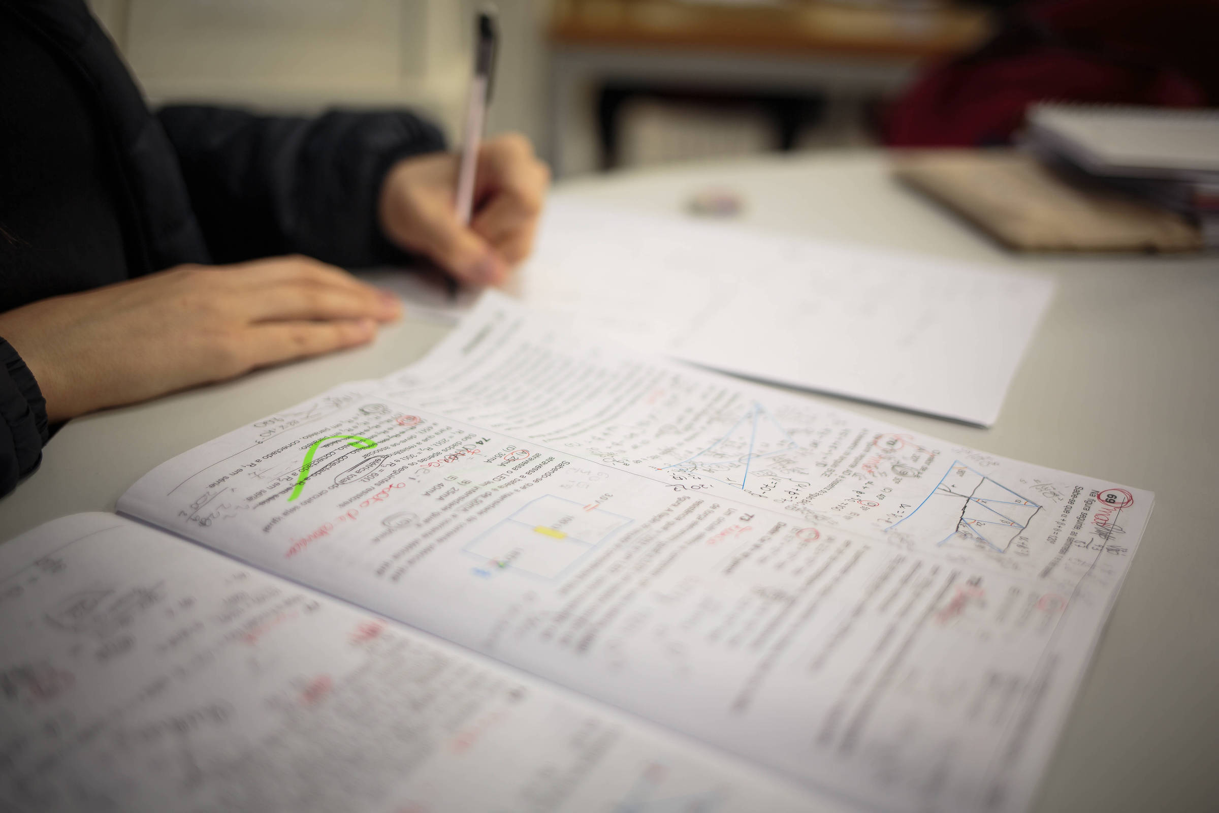 A imagem mostra uma mesa de estudo com um caderno aberto, onde há anotações e gráficos desenhados. Uma mão está segurando uma caneta, enquanto a outra parte do caderno apresenta textos e desenhos em caneta colorida. Ao fundo, há folhas em branco e outros materiais de estudo.