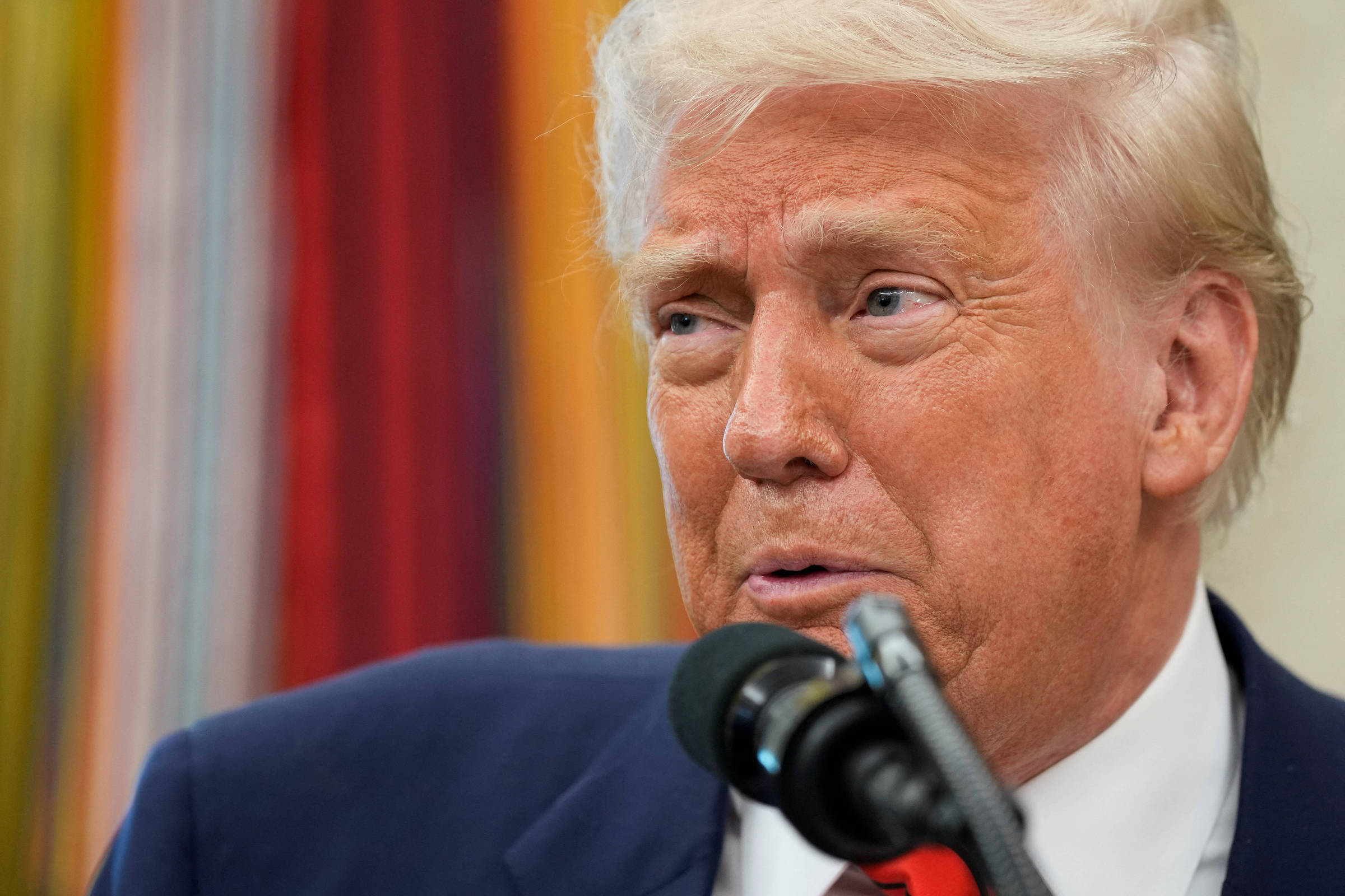 Um homem com cabelo loiro e pele clara está falando em um microfone. Ele usa um terno escuro e parece estar em um evento formal. Ao fundo, há uma parede com cores vibrantes e listras. O homem tem uma expressão facial que sugere que está em um momento de reflexão ou consideração.