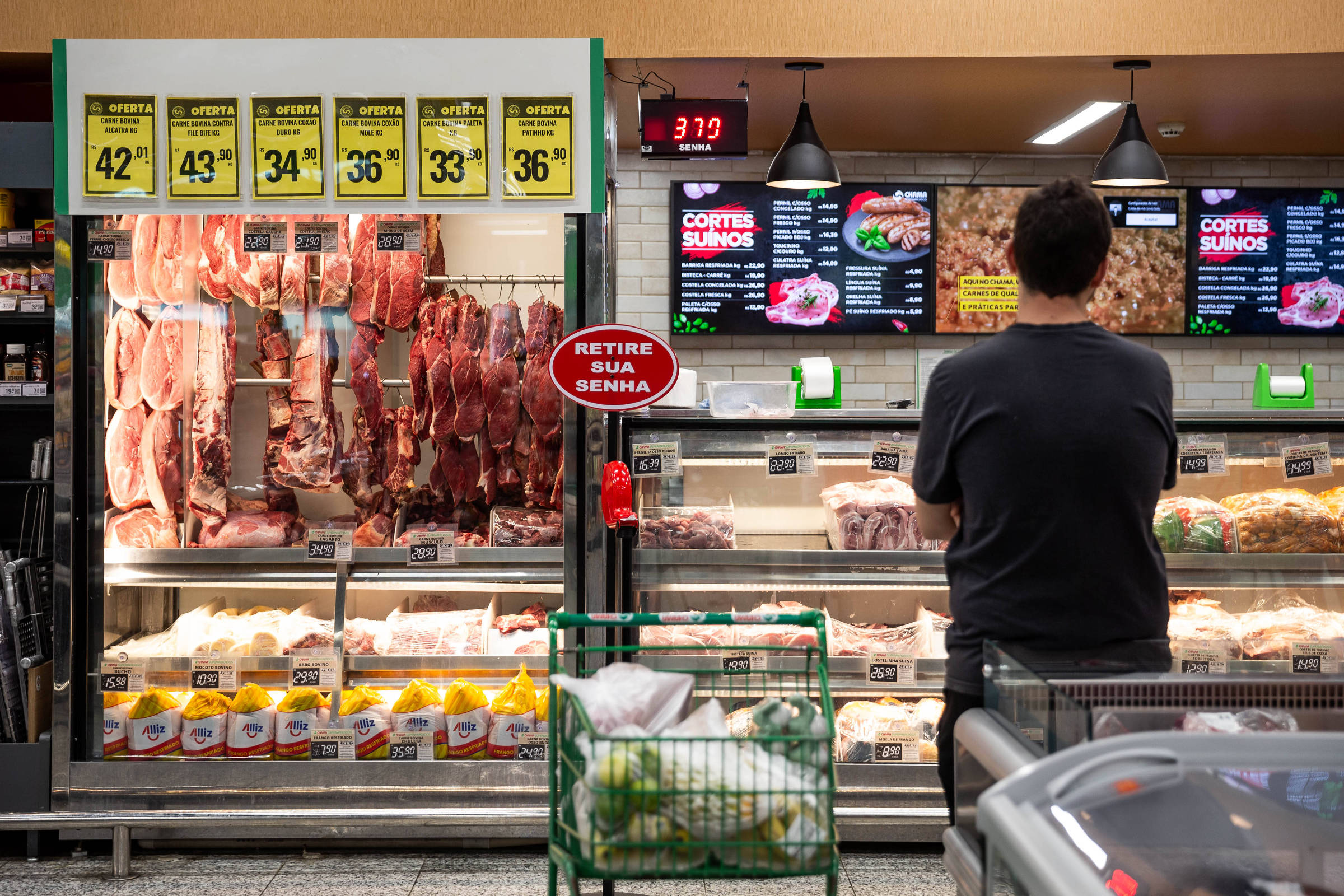 A imagem mostra um cliente em um supermercado, de costas, observando a seção de carnes. À esquerda, há um balcão refrigerado com várias peças de carne penduradas e embaladas, com preços visíveis. Acima do balcão, há placas com preços de diferentes cortes. À direita, um painel digital exibe informações sobre cortes de carne suína. Um carrinho de compras verde está ao lado do cliente, que parece estar esperando para ser atendido.