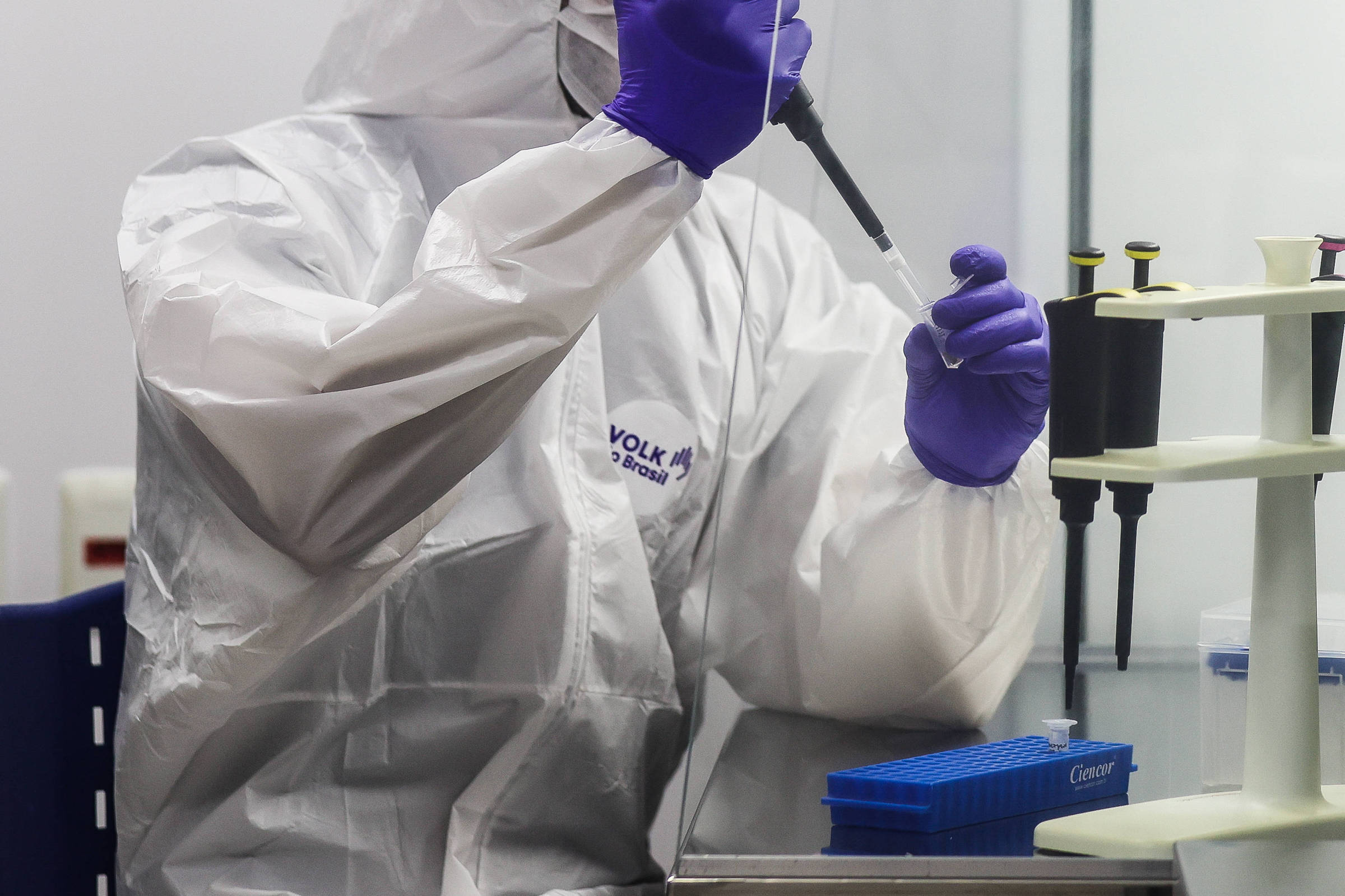 Um profissional de laboratório está usando um pipetador em um ambiente controlado. Ele está vestido com um traje de proteção completo, incluindo uma máscara e luvas roxas. Ao fundo, há uma bancada com equipamentos de laboratório e recipientes.