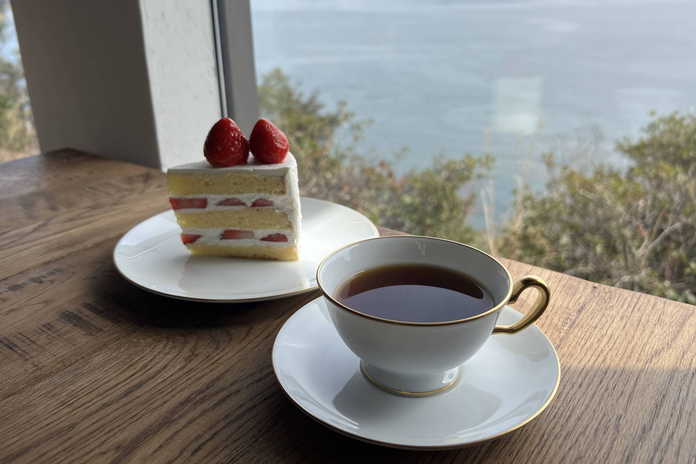 A imagem mostra um pedaço de bolo decorado com morangos em um prato branco, ao lado de uma xícara de café em uma mesa de madeira. Ao fundo, há uma vista de um lago ou mar com ilhas ao longe, sob um céu claro.