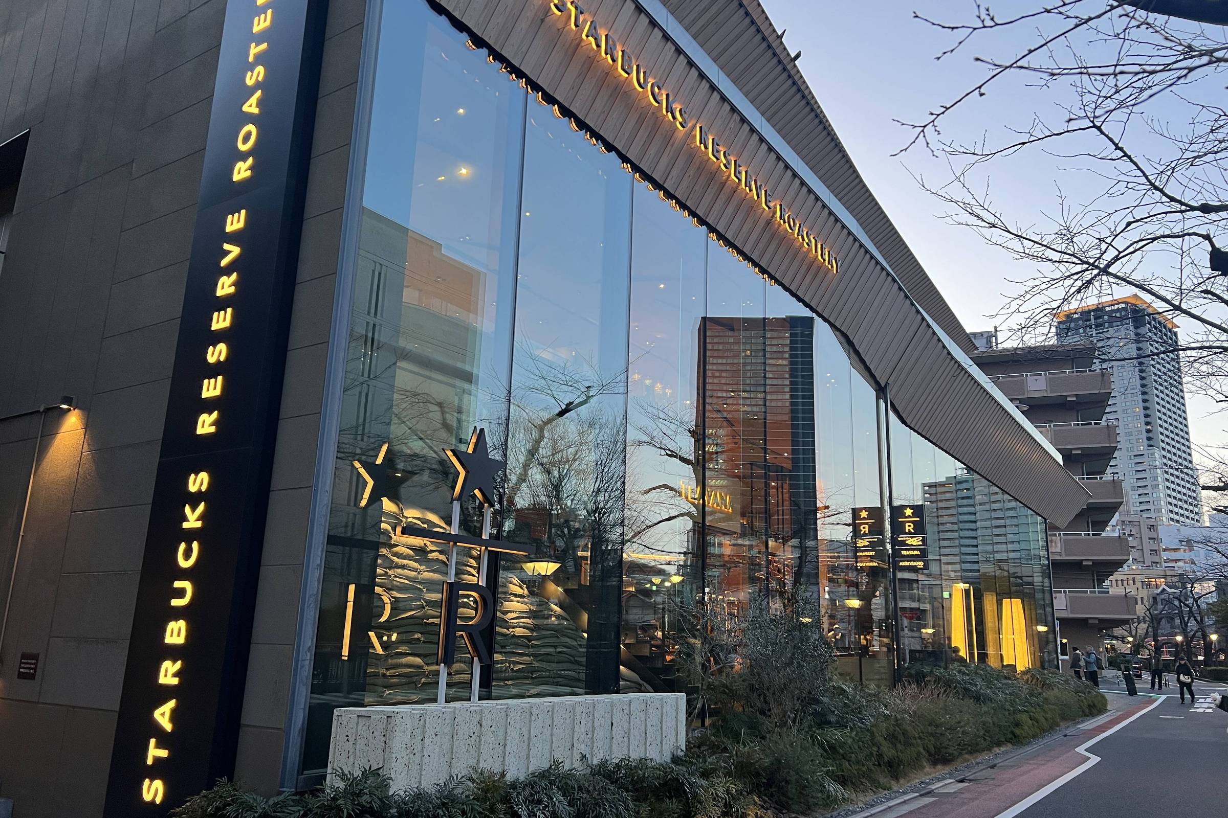 A imagem mostra a fachada de um Starbucks Reserve Roastery, com grandes janelas de vidro refletindo o céu e a cidade ao redor. O nome