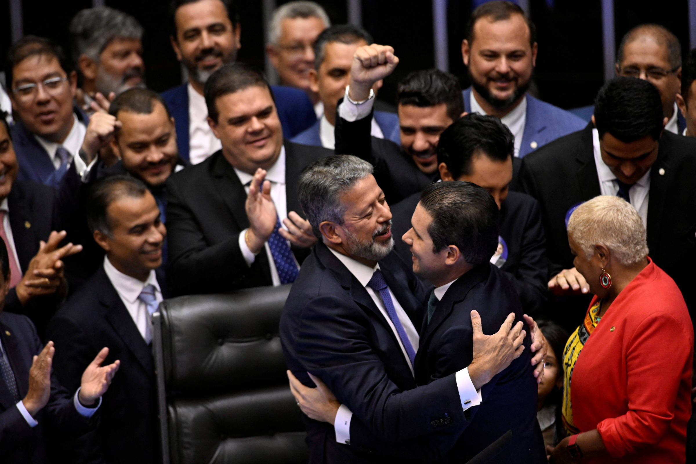 A imagem mostra um grupo de homens e mulheres em um ambiente de congresso, aparentemente celebrando um momento significativo. No centro, dois homens se abraçam, enquanto outros ao redor aplaudem e levantam os braços em sinal de comemoração. A maioria das pessoas está vestida formalmente, com ternos e gravatas. Há uma atmosfera de alegria e camaradagem entre os presentes.