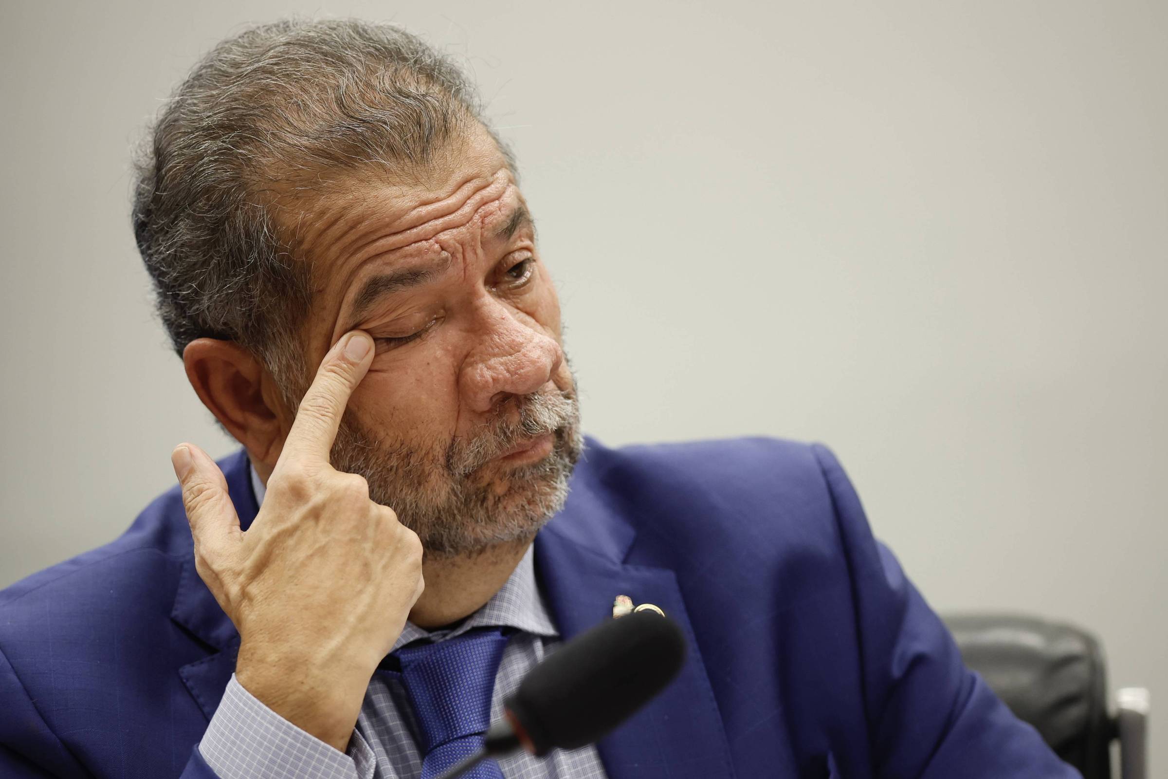 Um homem com cabelo grisalho e barba, vestindo um terno azul e uma camisa clara, está sentado em uma mesa. Ele está tocando seu rosto com a mão, parecendo pensativo ou reflexivo. O fundo é neutro e desfocado, sugerindo um ambiente formal, possivelmente uma reunião ou audiência.