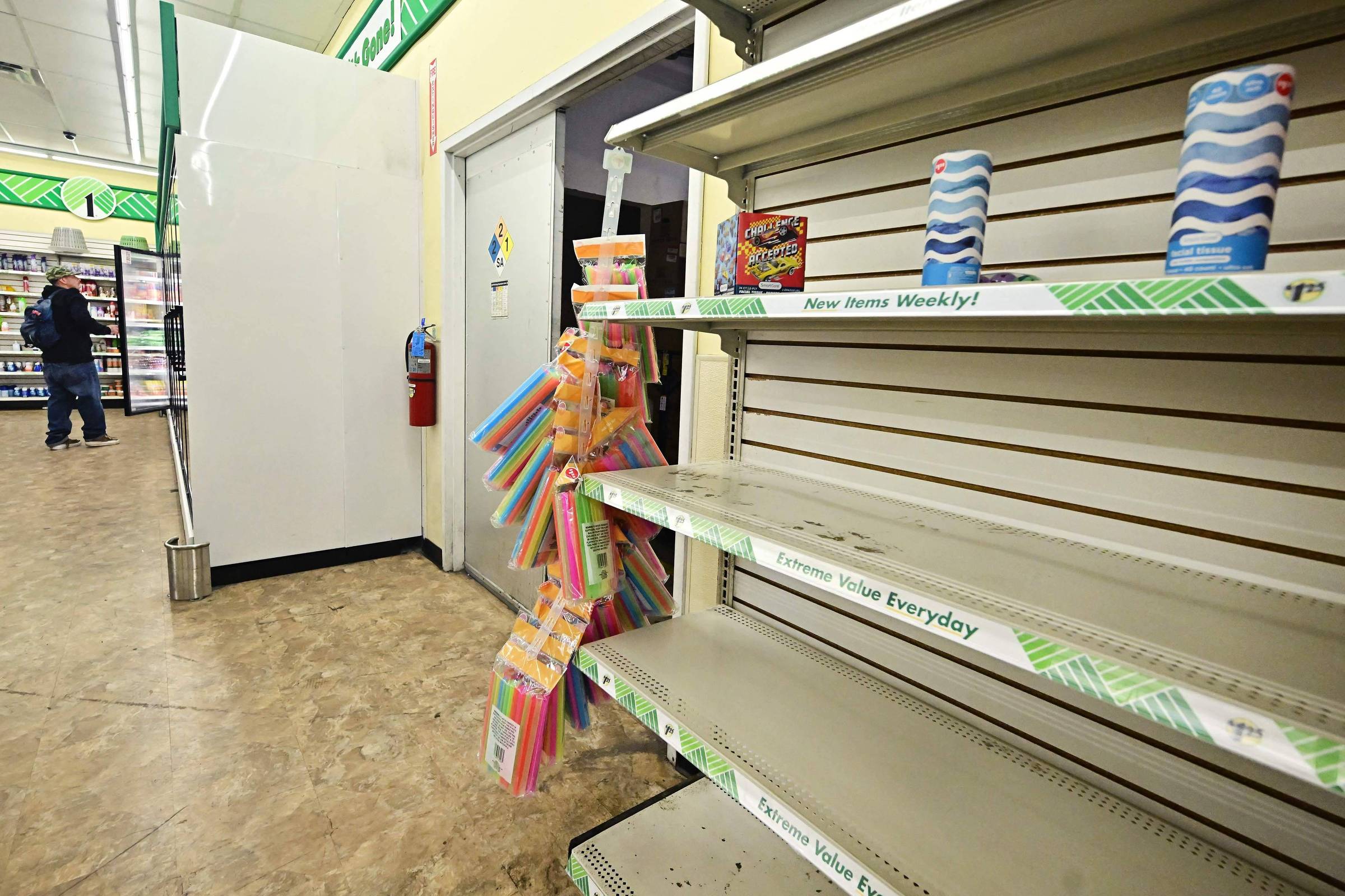 A imagem mostra prateleiras vazias em uma loja, com algumas mercadorias expostas. À esquerda, há um cliente de costas, e à direita, um suporte com produtos coloridos, como canetas e outros itens de papelaria. O ambiente é bem iluminado, com paredes claras e um piso de linóleo.