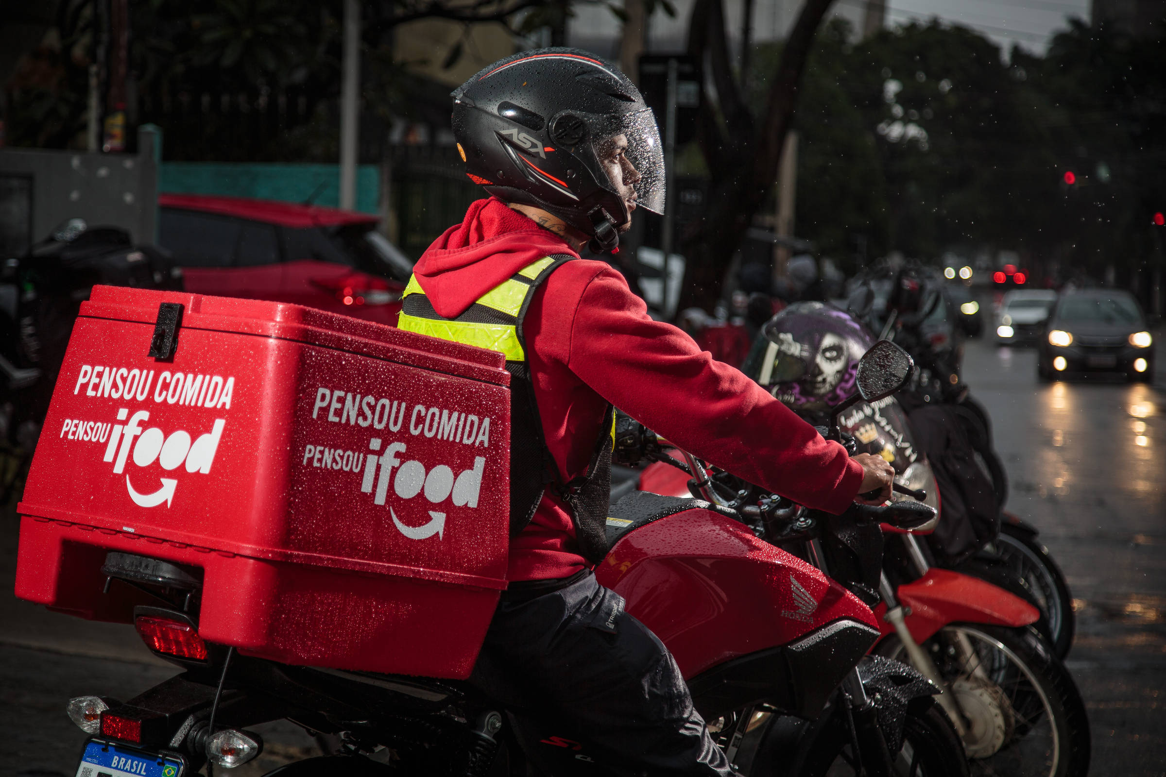 Um entregador de comida está montado em uma motocicleta vermelha, usando um capacete e uma jaqueta vermelha com detalhes em amarelo. Ele carrega uma caixa de entrega vermelha nas costas, com o logo da empresa iFood e a frase