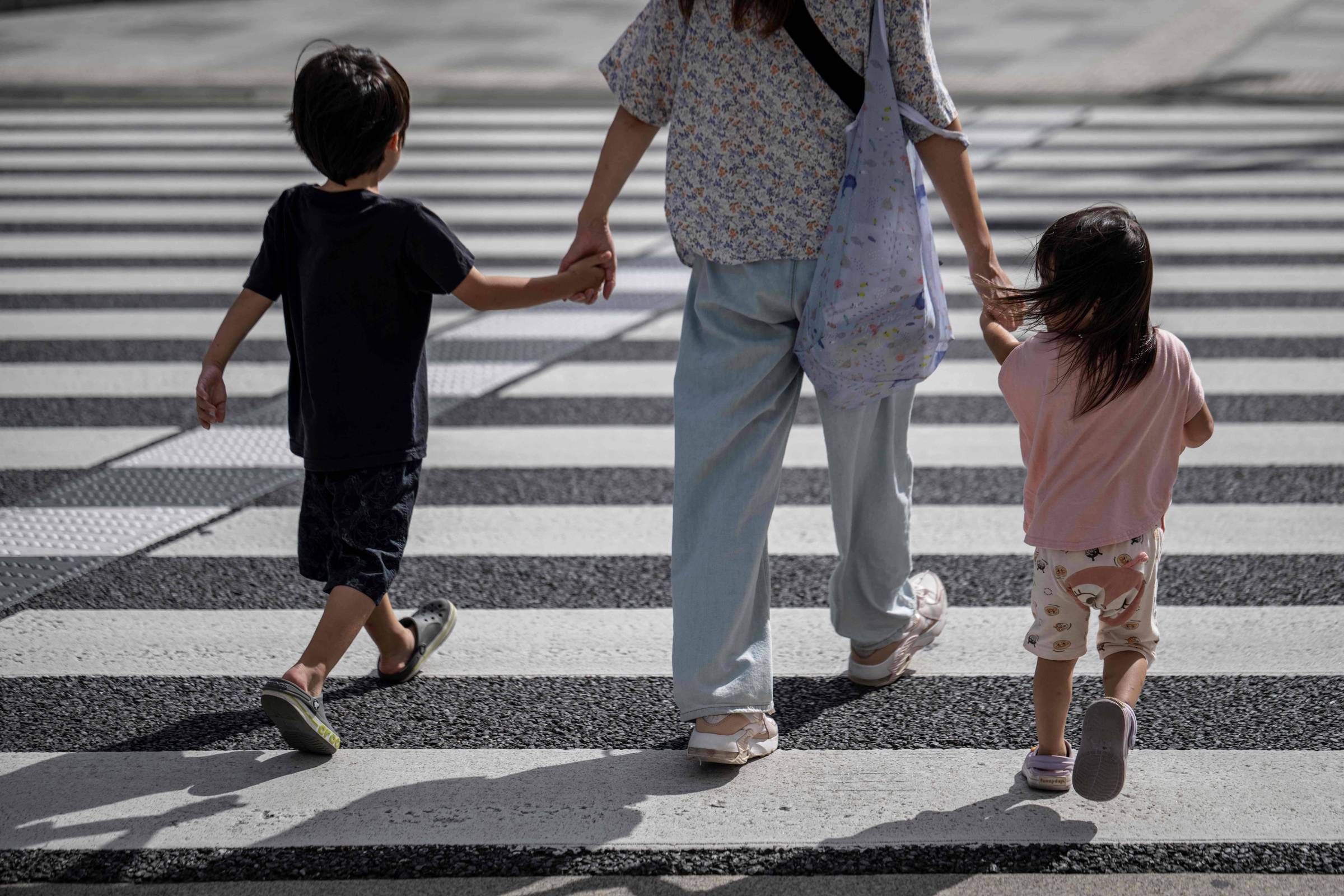 A imagem mostra uma mulher segurando as mãos de duas crianças enquanto atravessam uma faixa de pedestres. A mulher está vestindo uma blusa clara e calças largas, e carrega uma bolsa. As crianças, um menino e uma menina, estão usando roupas casuais. O menino está à esquerda, vestindo uma camiseta escura e shorts, enquanto a menina está à direita, usando uma camiseta rosa e calças claras.