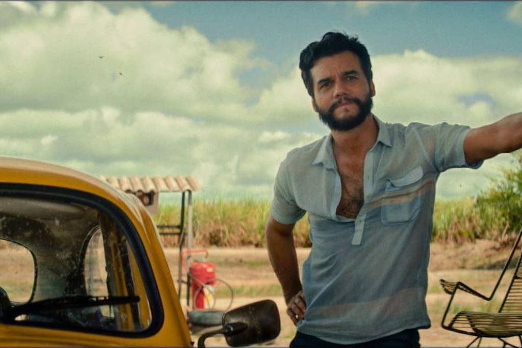 Um homem com barba e cabelo encaracolado está posicionado ao lado de um carro amarelo. Ele usa uma camisa de botão clara e está em um ambiente ao ar livre, com um céu azul e nuvens. Ao fundo, há canaviais e uma estrutura simples que parece ser uma garagem ou abrigo. O homem tem uma expressão relaxada e confiante.
