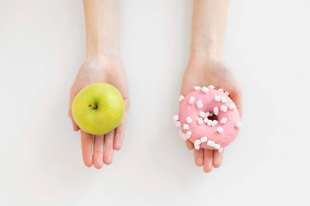 Uma mão segurando uma maçã e outra segurando uma rosquinha açúcarada cor de rosa do tipo donut
