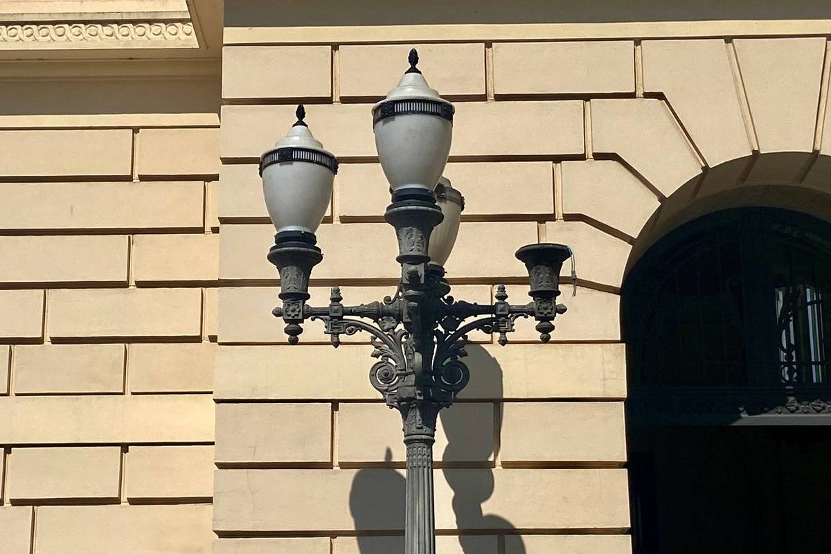A imagem mostra uma luminária de rua clássica, com três lâmpadas em um suporte ornamentado. A luminária é de metal escuro e está posicionada ao lado de uma parede de edifício de cor clara, que apresenta um padrão de pedras retangulares. Ao fundo, é visível uma porta com grades.