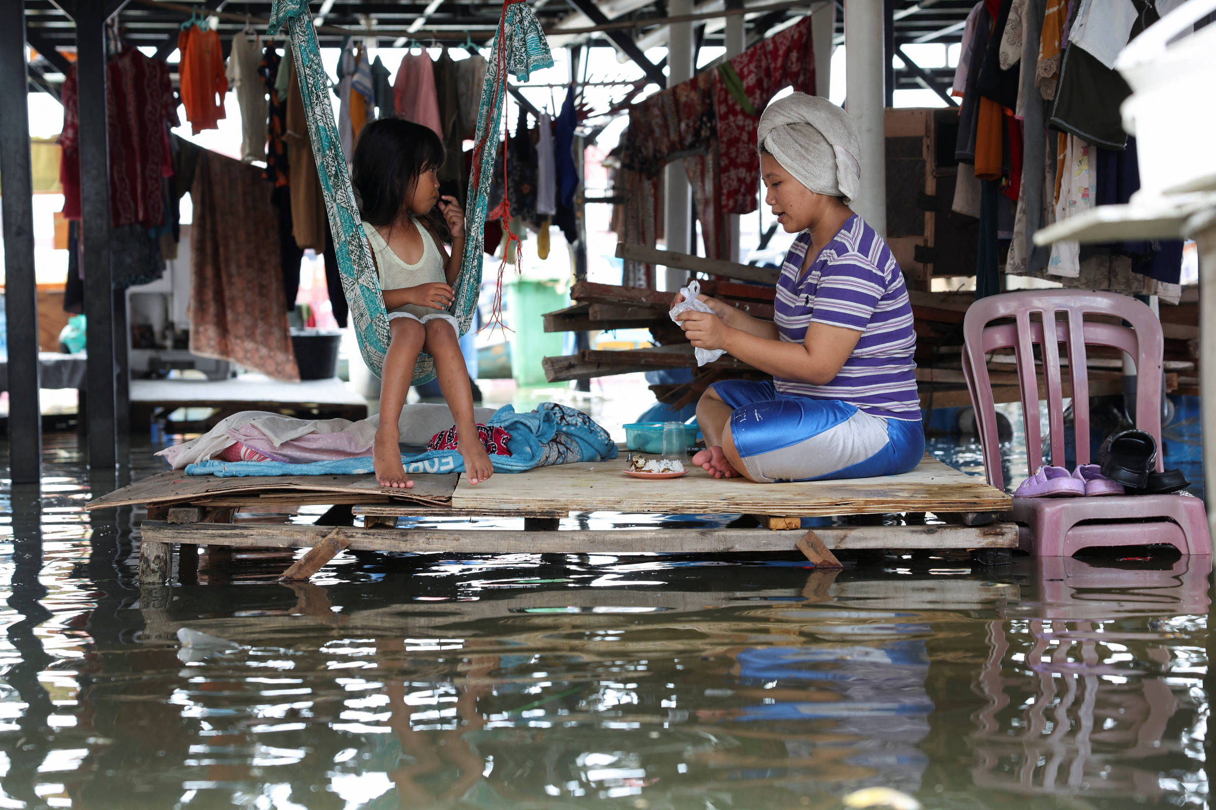A imagem mostra uma cena em uma área inundada, onde uma mulher senta em uma plataforma de madeira, enquanto uma menina pequena está sentada em uma rede. A água está visivelmente alta ao redor, e roupas estão penduradas em um varal próximo. A mulher parece estar ocupada com algo em suas mãos, enquanto a menina observa.