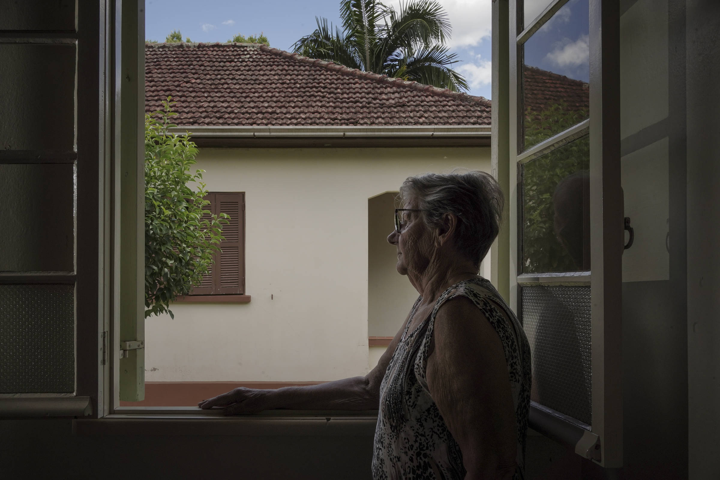 Uma mulher idosa está de pé ao lado de uma janela aberta, olhando para fora. Ela usa óculos e uma blusa de alças. A vista do lado de fora mostra uma casa com telhado de cerâmica e uma árvore verde. O céu está parcialmente nublado, e a luz natural ilumina a cena.