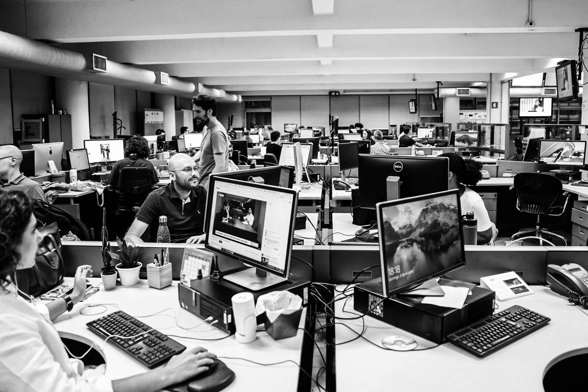 A imagem mostra um ambiente de escritório moderno, com várias mesas de trabalho e computadores. Há pessoas sentadas em algumas mesas, algumas trabalhando em seus computadores, enquanto outras interagem. O espaço é bem iluminado, com janelas ao fundo e várias telas de computador visíveis. Plantas decorativas estão presentes nas mesas, e há uma atmosfera de colaboração e atividade.