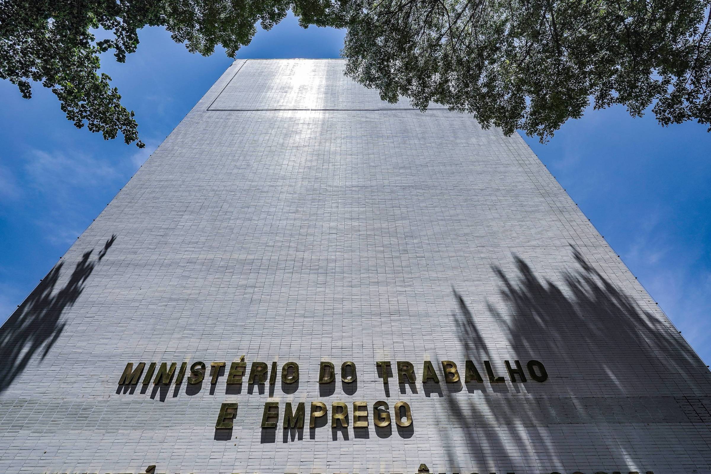 A imagem mostra a fachada de um edifício alto, que é o Ministério do Trabalho e Emprego. O nome do ministério está escrito em letras grandes e douradas na parte inferior da parede do edifício. O céu está claro e azul, e há algumas árvores na parte superior da imagem, que projetam sombras na parede do edifício