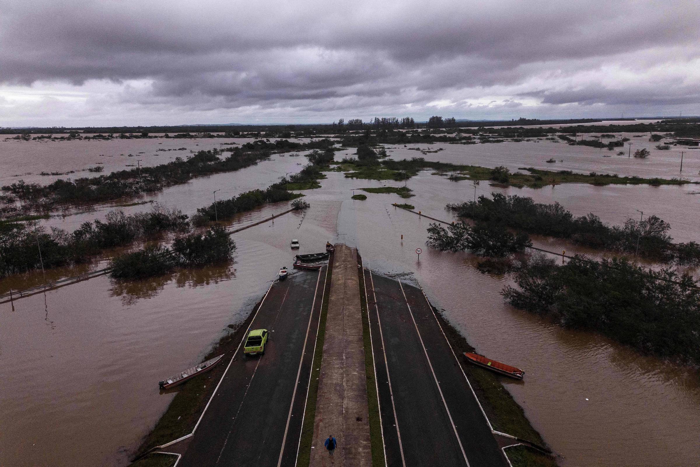 A imagem mostra uma área urbana severamente inundada, com grandes extensões de água cobrindo a vegetação e as estradas. No centro, há uma estrada que se estende em direção a um ponto onde a água parece mais profunda. Algumas árvores e arbustos são visíveis, mas a maior parte da paisagem está submersa. O céu está nublado, indicando condições climáticas adversas.