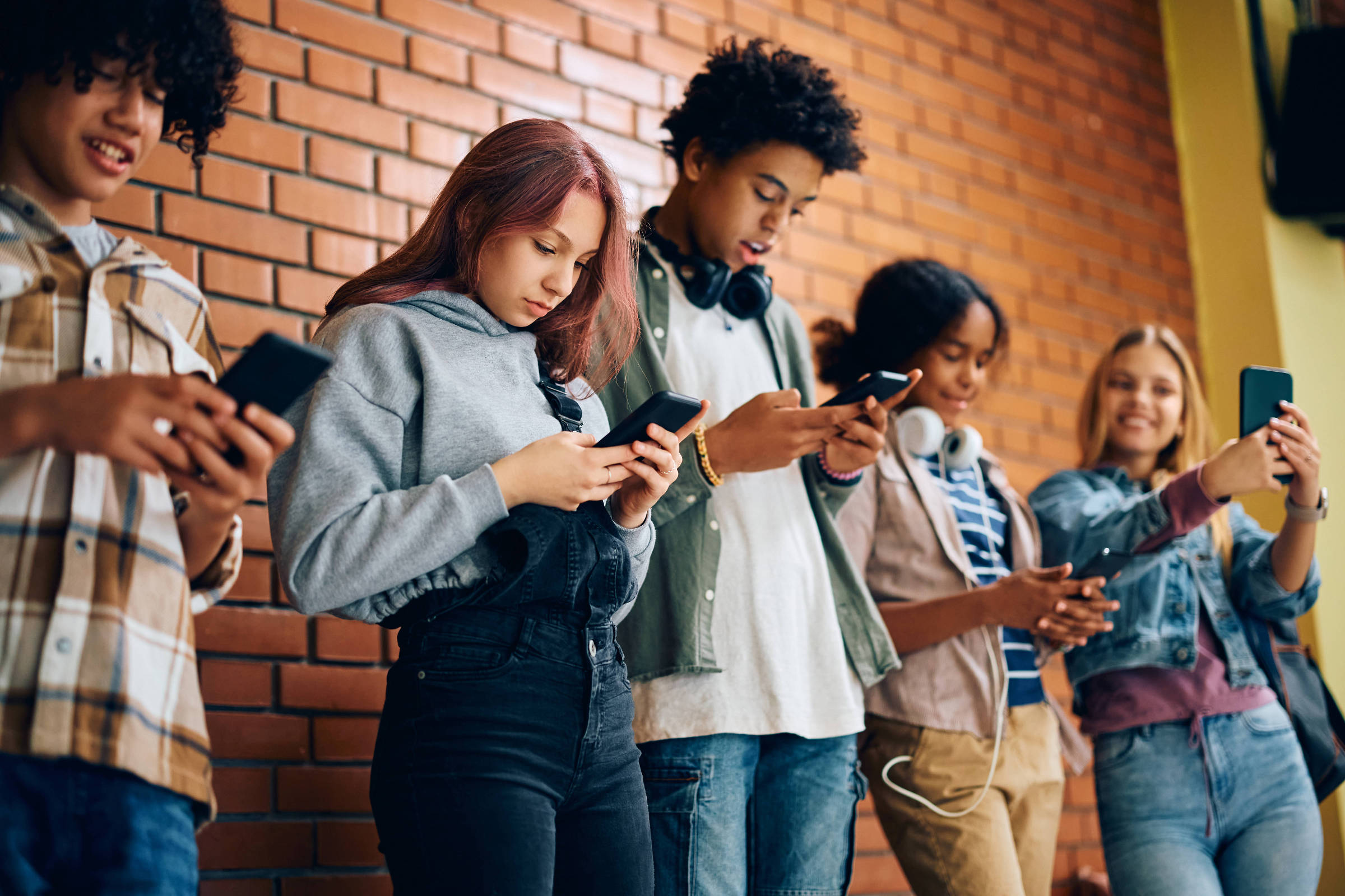 A imagem mostra um grupo de cinco jovens em pé, alinhados contra uma parede de tijolos. Todos estão olhando para seus smartphones. A jovem no centro tem cabelo longo e liso, vestindo um moletom cinza e calças escuras. Os outros jovens estão usando diferentes estilos de roupas casuais e também seguram seus celulares. O ambiente parece ser interno, com uma iluminação suave.