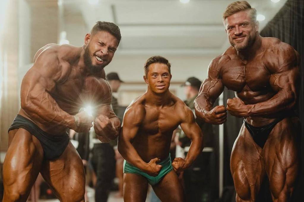 A imagem mostra três fisiculturistas posando juntos. Dois deles são adultos musculosos, um à esquerda e outro à direita, ambos com pele bronzeada e exibindo seus músculos. O fisiculturista no centro é menor em estatura, mas também musculoso, usando um sunga verde. O fundo é desfocado, mas parece ser um ambiente de competição, com iluminação suave e um tapete marrom no chão.
