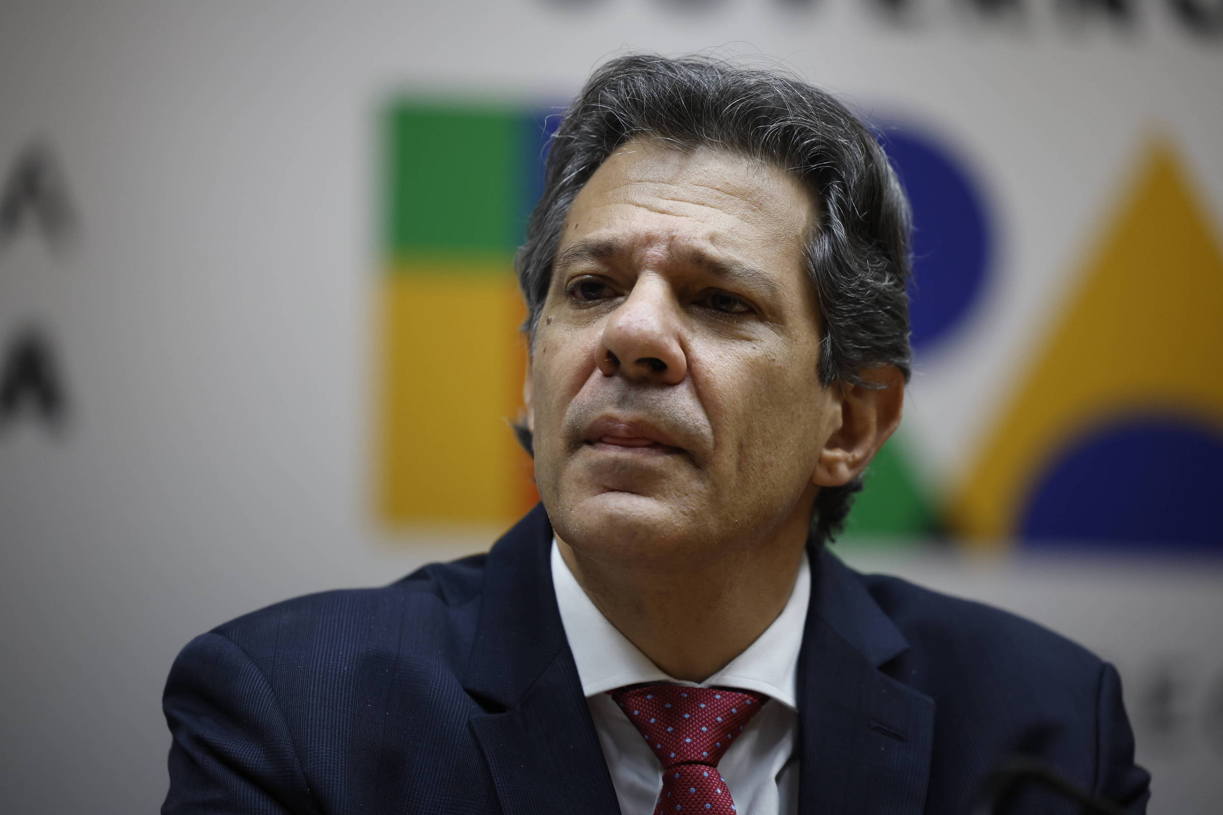 Um homem com cabelo grisalho e liso, usando um terno escuro e uma gravata vermelha, está sentado em uma mesa. Ele parece pensativo e está olhando para frente, com uma expressão séria. Ao fundo, há um painel com cores verde, azul e amarelo, que sugere um ambiente de conferência ou coletiva de imprensa.