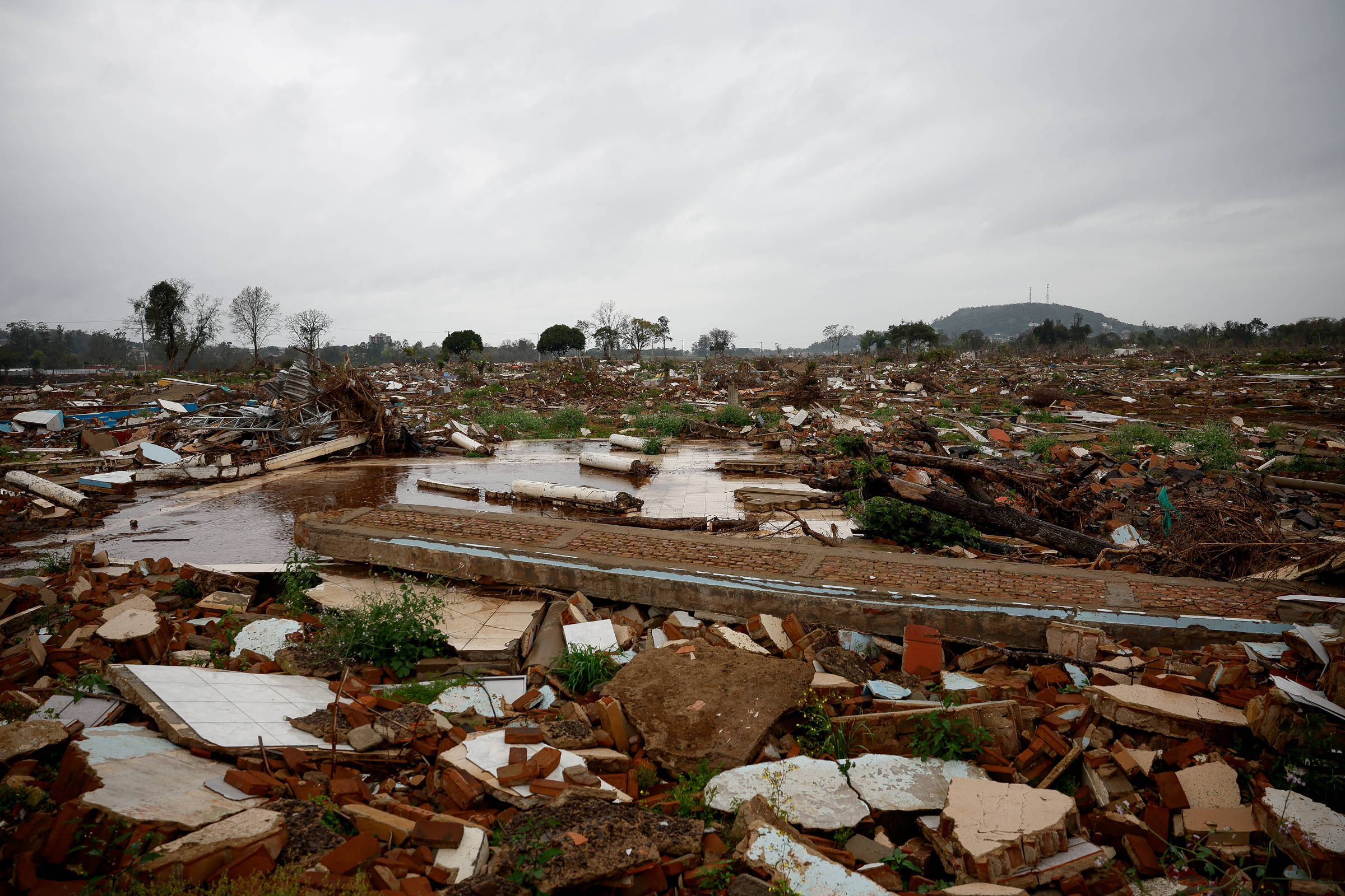 A imagem mostra uma área devastada, com destroços de madeira e telhas espalhados pelo chão. Há uma grande quantidade de entulho e restos de construções, com algumas áreas alagadas. O céu está nublado, sugerindo um clima sombrio, e ao fundo é possível ver uma colina e algumas árvores.