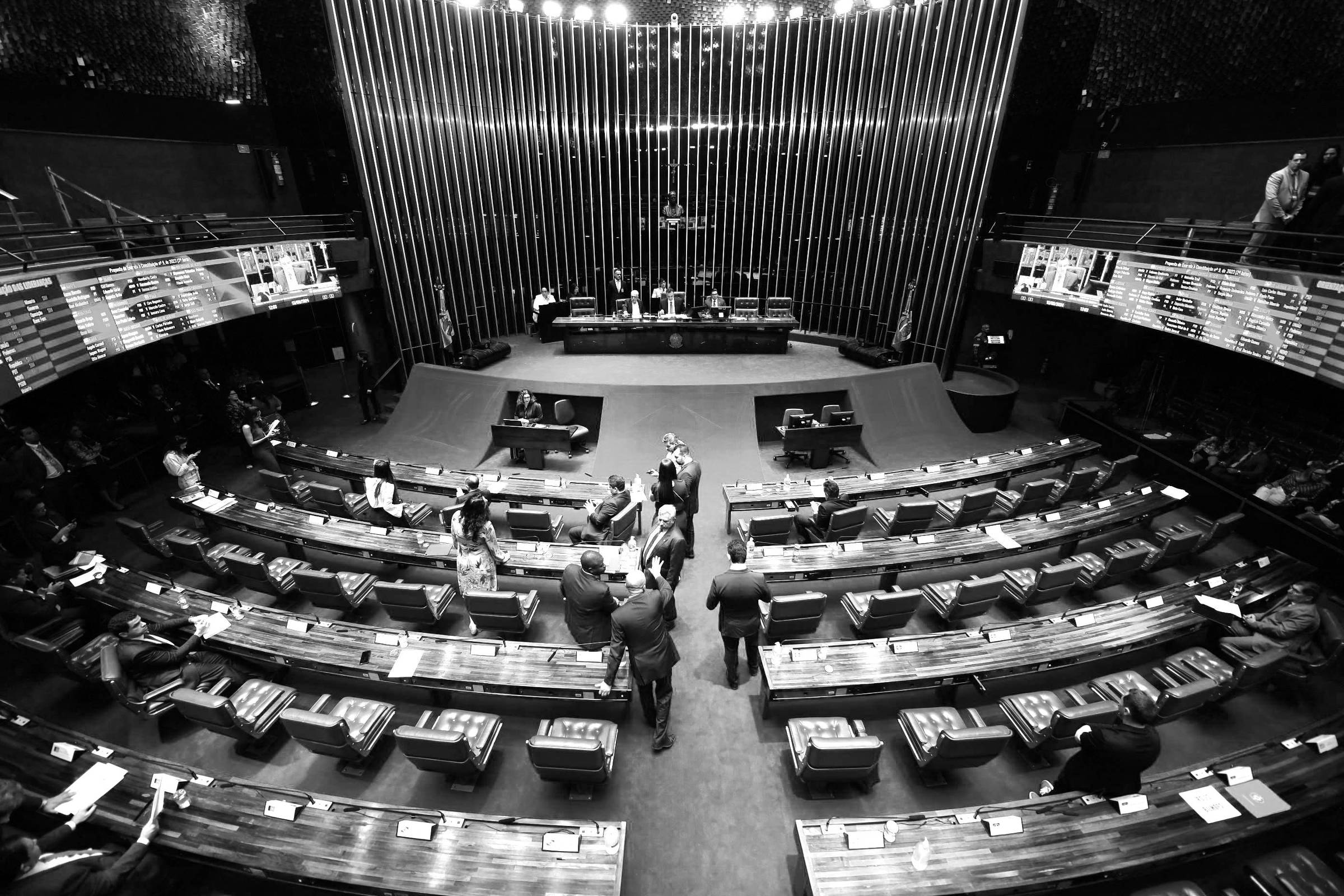 A imagem mostra o interior do plenário do Senado Federal do Brasil. O ambiente é amplo, com cadeiras azuis dispostas em fileiras e mesas de madeira. Ao fundo, há uma mesa de autoridades e telas eletrônicas exibindo informações. O chão é coberto por um tapete azul e há algumas pessoas circulando pelo espaço.