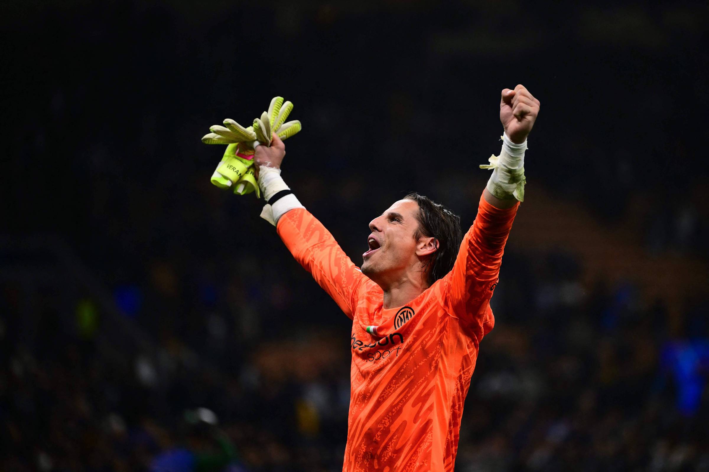 Um goleiro vestindo um uniforme laranja e luvas de goleiro levanta os braços em celebração. Ele parece estar em um estádio, com uma multidão ao fundo, e expressa alegria e emoção. O ambiente é iluminado, sugerindo um momento importante em uma partida de futebol.