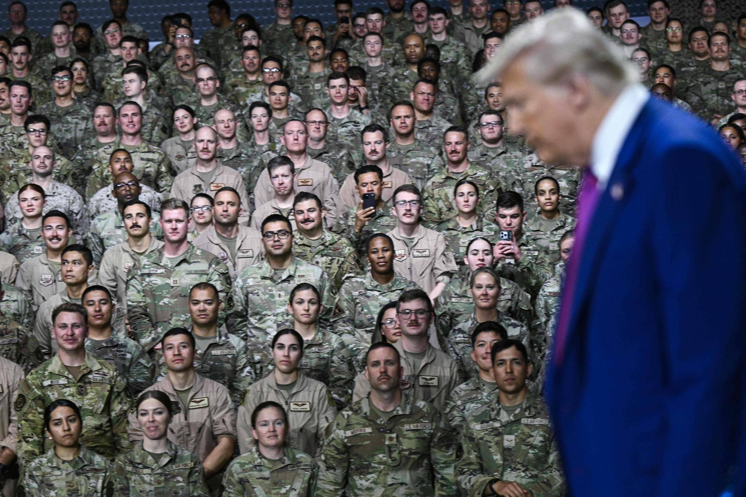 A imagem mostra um grande grupo de soldados em uniformes militares, organizados em várias fileiras. Eles estão olhando para a câmera, com expressões neutras. Na frente, um homem de terno azul e gravata rosa está se afastando, parcialmente fora de foco. O fundo é uma parede com um padrão discreto.