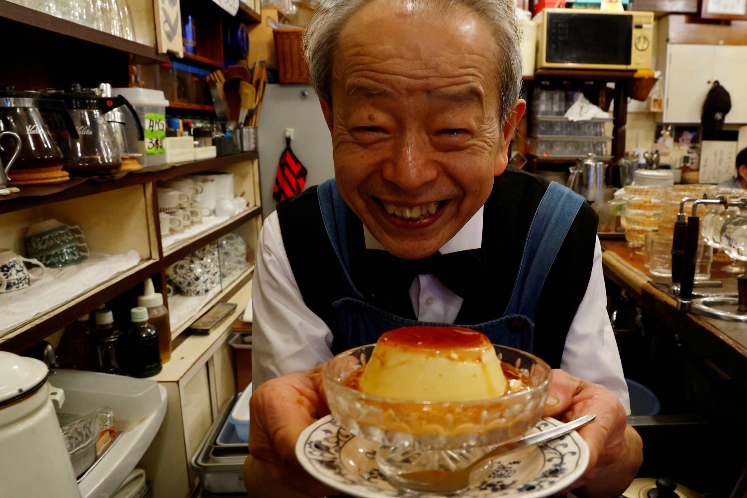 Um homem idoso, sorridente, está segurando uma sobremesa em uma tigela de vidro. Ele está vestido com um colete azul e uma camisa branca, e está em um ambiente de café ou restaurante, com prateleiras cheias de utensílios e copos ao fundo. A sobremesa parece ser um pudim, com uma camada de caramelo por cima.