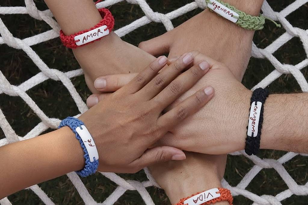 Cinco mãos estão sobrepostas em uma rede, cada uma usando uma pulseira colorida. As pulseiras têm etiquetas brancas com as palavras