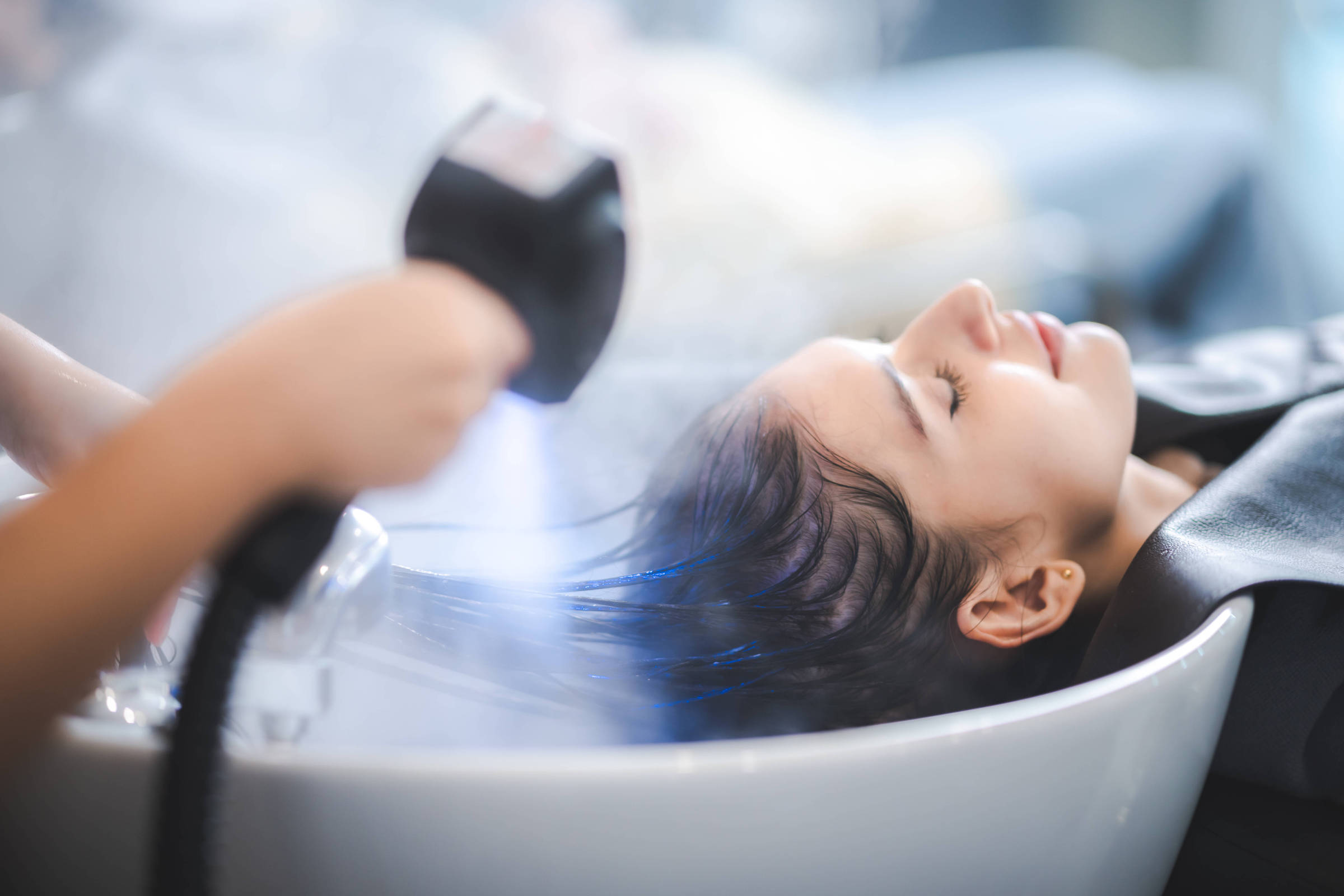 Uma mulher está deitada em uma pia de salão de beleza, recebendo um tratamento capilar. Ela tem os olhos fechados e parece relaxada. Uma pessoa está usando um aparelho para aplicar vapor ou um produto em seu cabelo, que está molhado e escorrendo na pia. O ambiente é iluminado e parece ser um salão moderno.