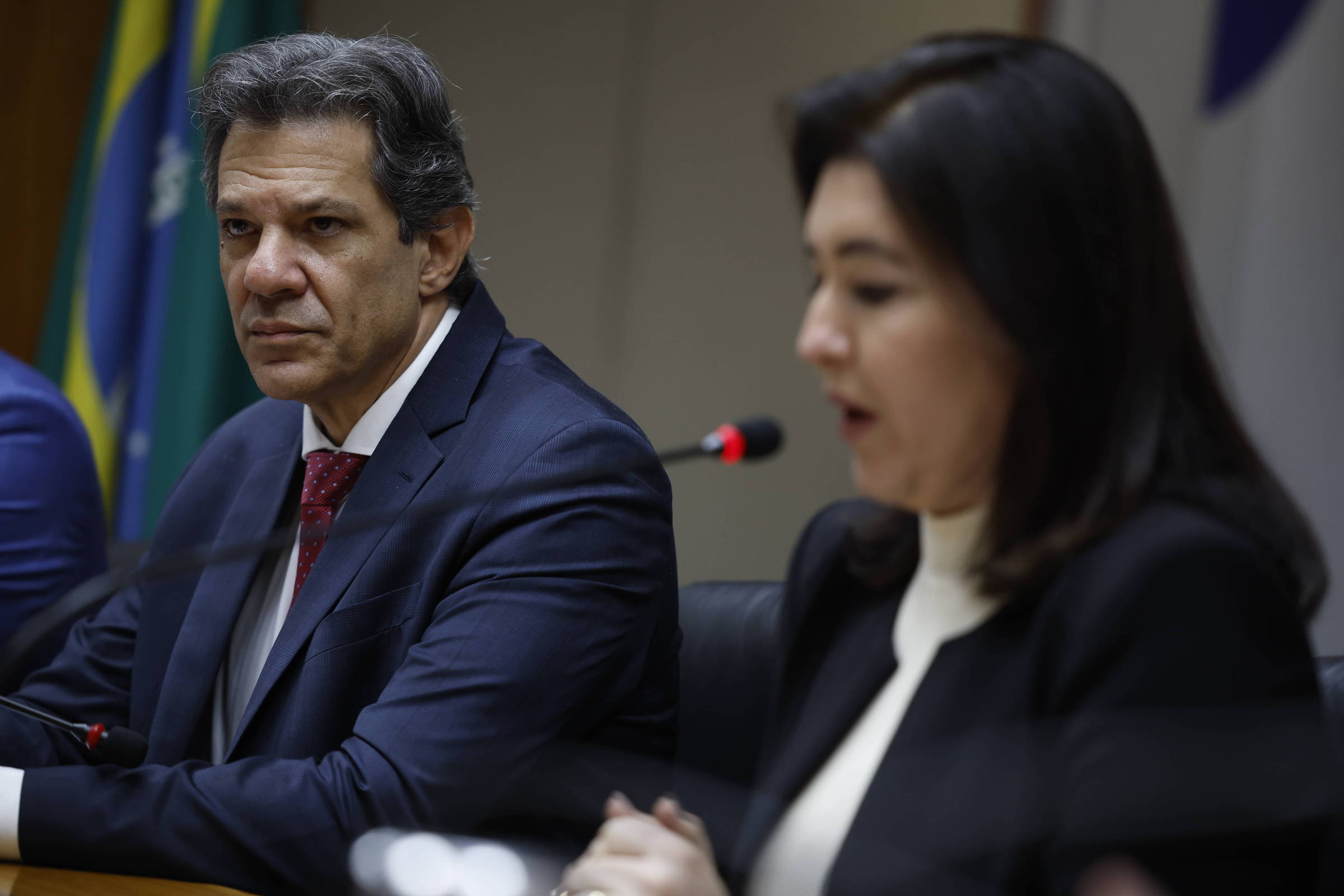A imagem mostra um homem e uma mulher em uma reunião. O homem, à esquerda, está usando um terno escuro e uma gravata vermelha, com uma expressão séria, olhando para a mulher à sua direita, que está falando. Ela usa um blazer preto e uma blusa branca, e está posicionada em frente a um microfone. Ao fundo, há bandeiras do Brasil e outros elementos de um ambiente formal de reunião.