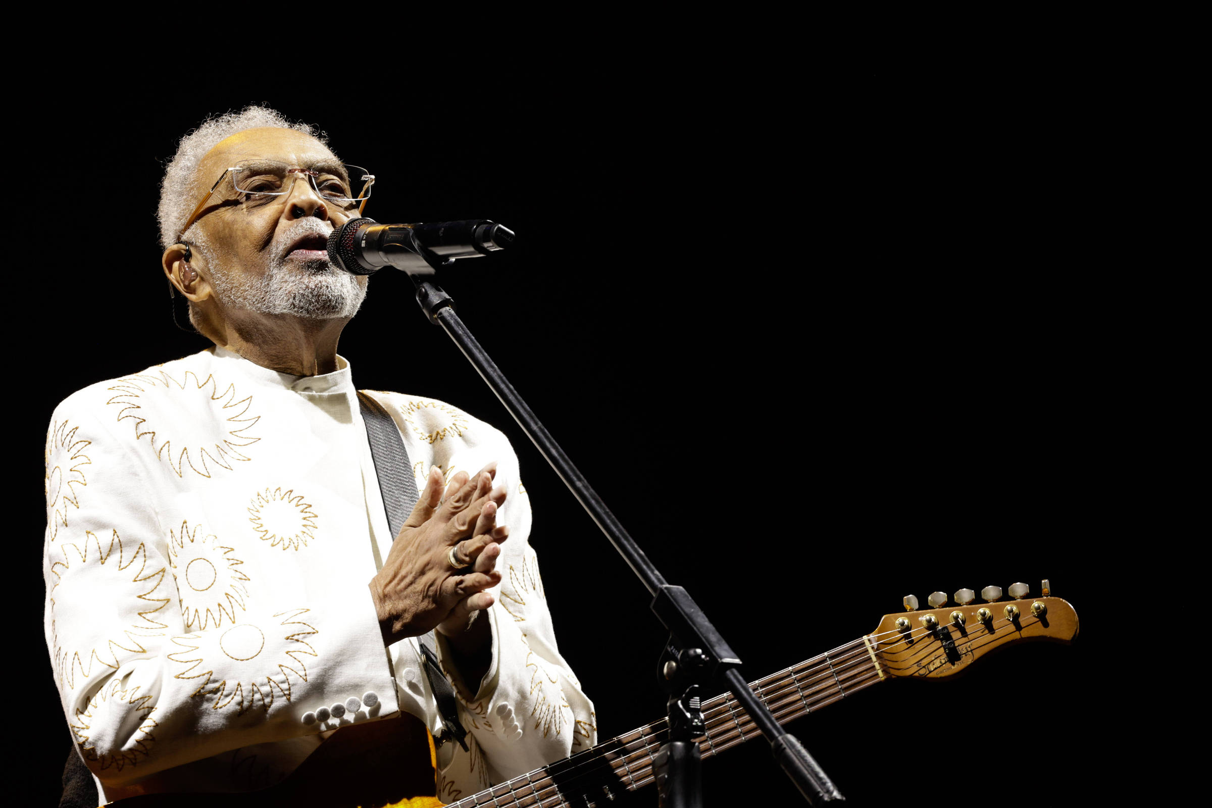 Um artista está se apresentando em um palco, segurando um microfone e uma guitarra. Ele usa uma roupa branca com detalhes em dourado e óculos. O fundo é escuro, destacando a figura do artista.