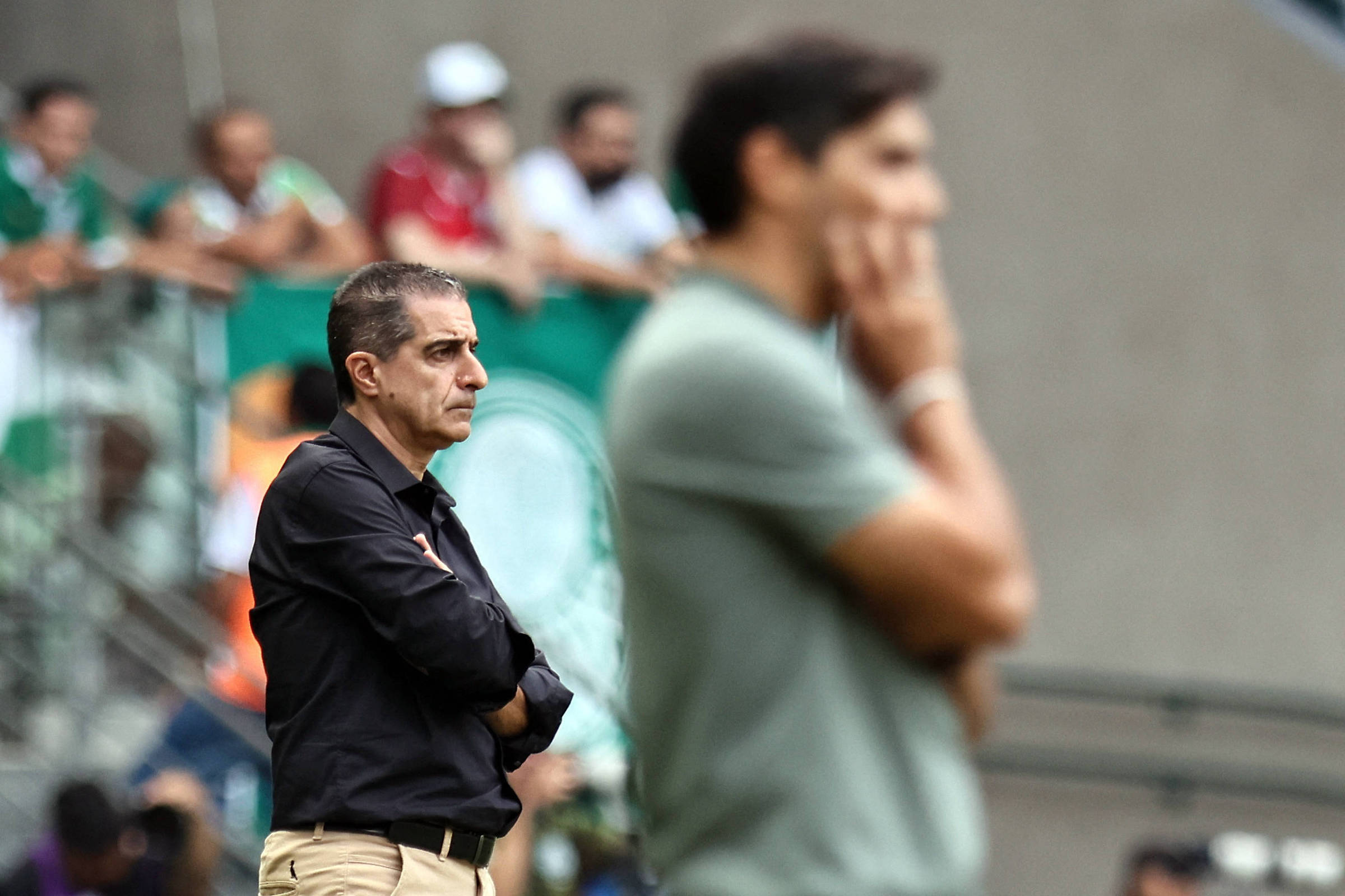 A imagem mostra dois técnicos de futebol em um campo. O técnico à esquerda está vestido com uma camisa preta e calças claras, enquanto o técnico à direita usa uma camiseta verde e calças escuras. Ao fundo, há torcedores visíveis, com bandeiras e camisas de times. A cena parece ser de um momento de tensão durante uma partida.