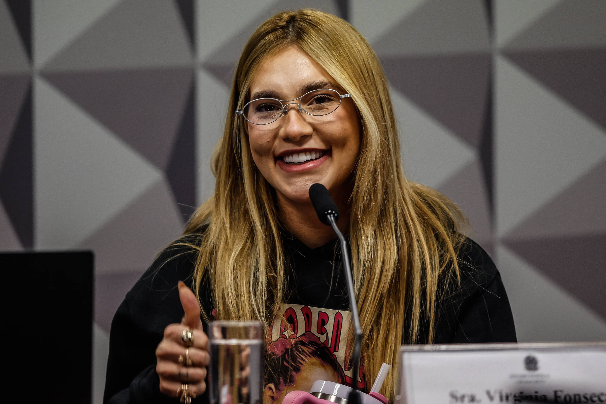Uma mulher com cabelo longo e liso, usando óculos e uma blusa preta, está sorrindo e fazendo um sinal de positivo com a mão. Ela está sentada em uma mesa com um copo de água e um microfone à sua frente. O fundo apresenta um padrão geométrico em tons de cinza.