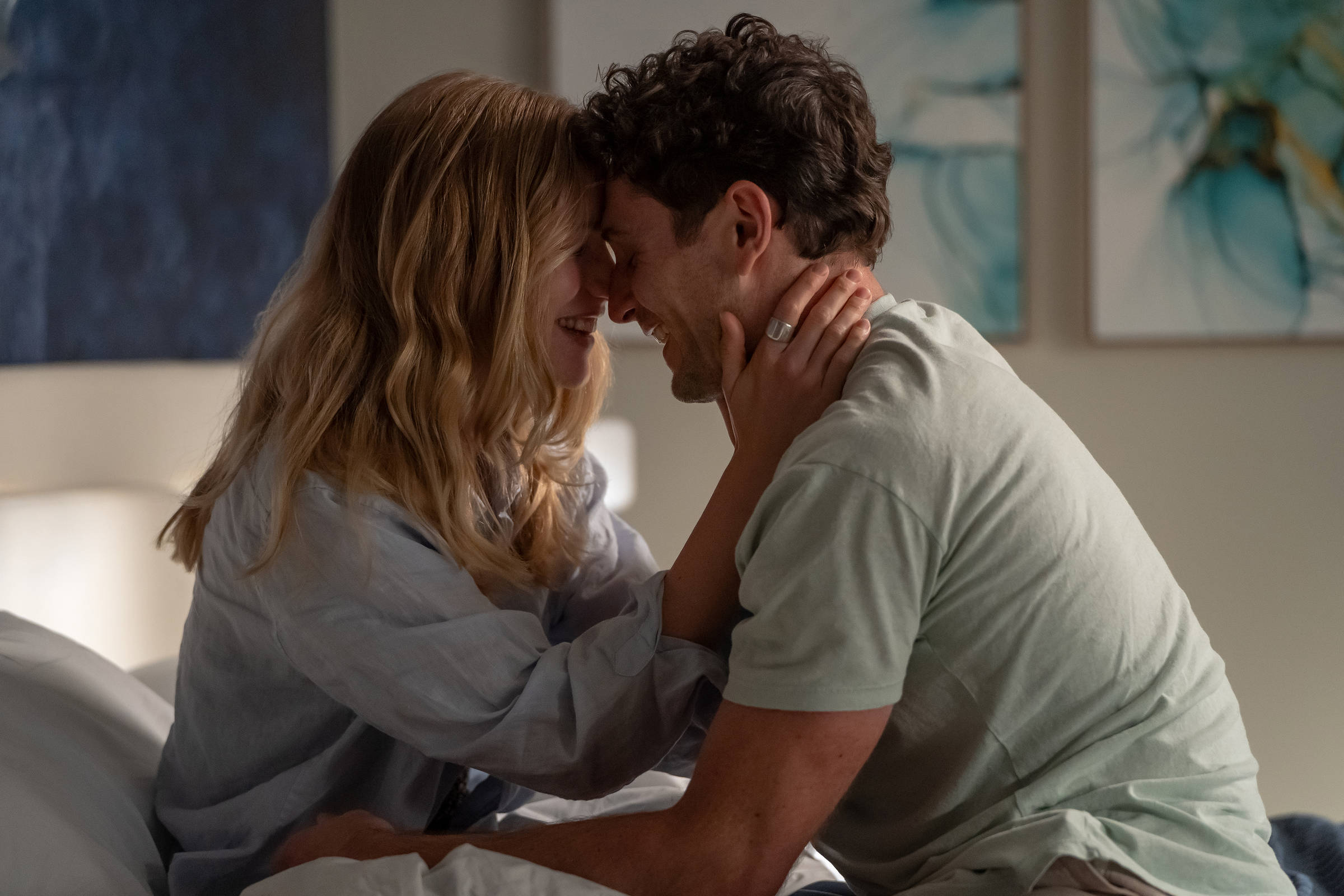 A imagem mostra um casal sentado em uma cama, com os rostos próximos e sorrindo um para o outro. A mulher tem cabelos longos e ondulados, e está vestindo uma camisa de botão clara. O homem tem cabelos curtos e está usando uma camiseta de cor clara. Eles estão em um ambiente bem iluminado, com quadros decorativos nas paredes ao fundo.