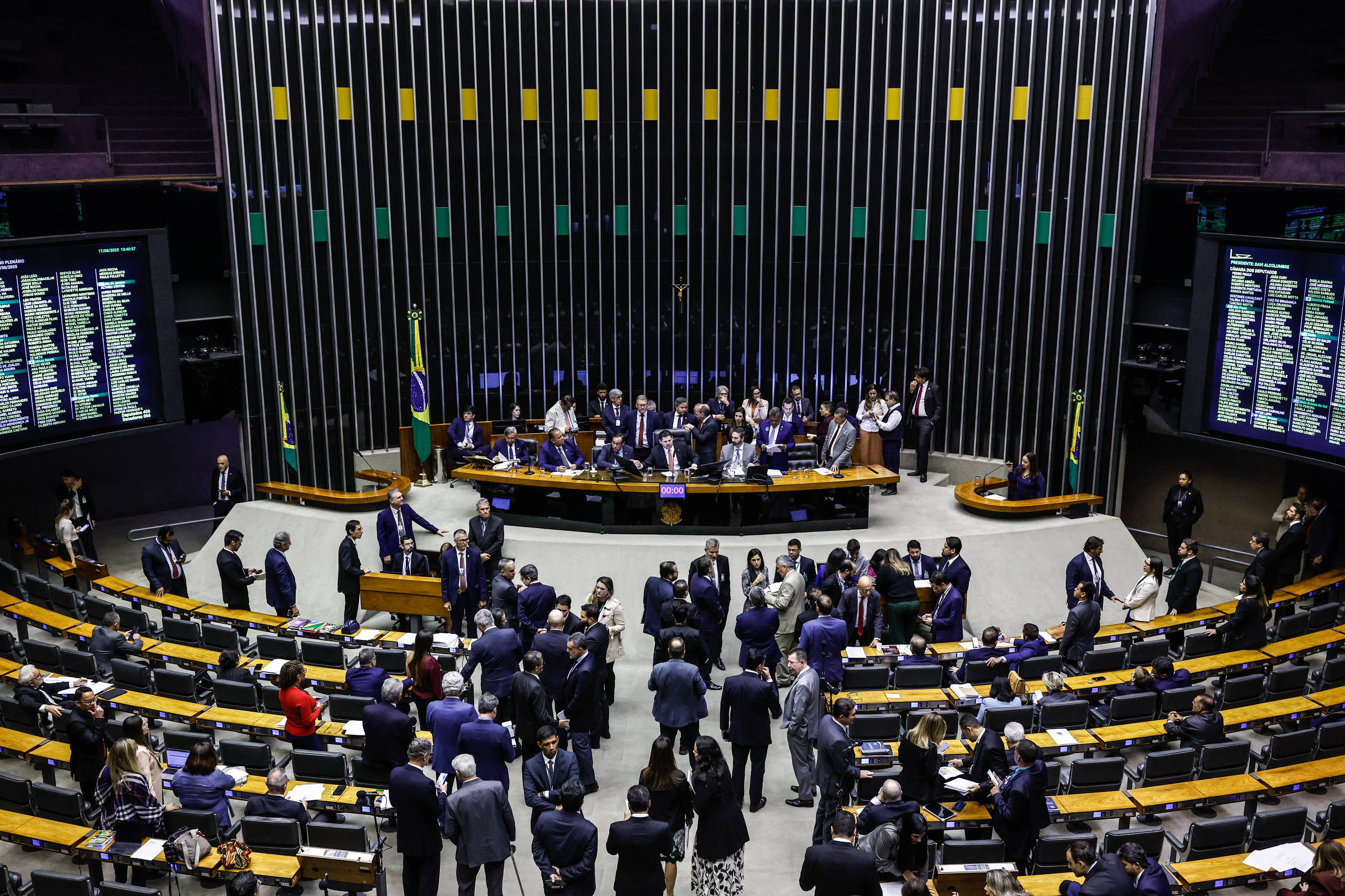 A imagem mostra o plenário da Câmara dos Deputados do Brasil, com várias pessoas em trajes formais interagindo. No fundo, há uma mesa com deputados e bandeiras do Brasil. As paredes são decoradas com painéis e telas que exibem informações. O ambiente é bem iluminado e moderno, com assentos dispostos em fileiras.