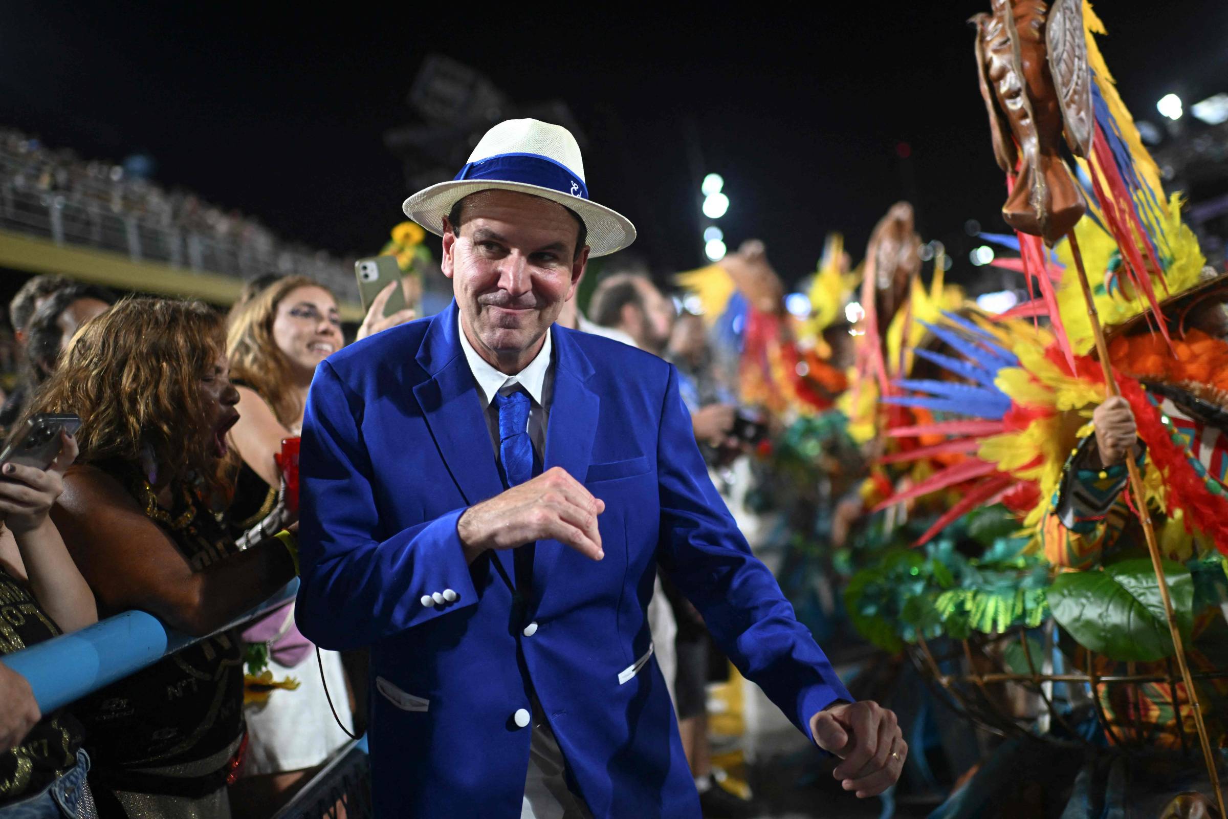 A imagem mostra um homem vestido com um terno azul e um chapéu branco, dançando e sorrindo em meio a uma multidão durante um evento de carnaval. Ao fundo, há pessoas vestidas com fantasias coloridas e plumas, criando um ambiente festivo. A iluminação é intensa, destacando a alegria do momento.