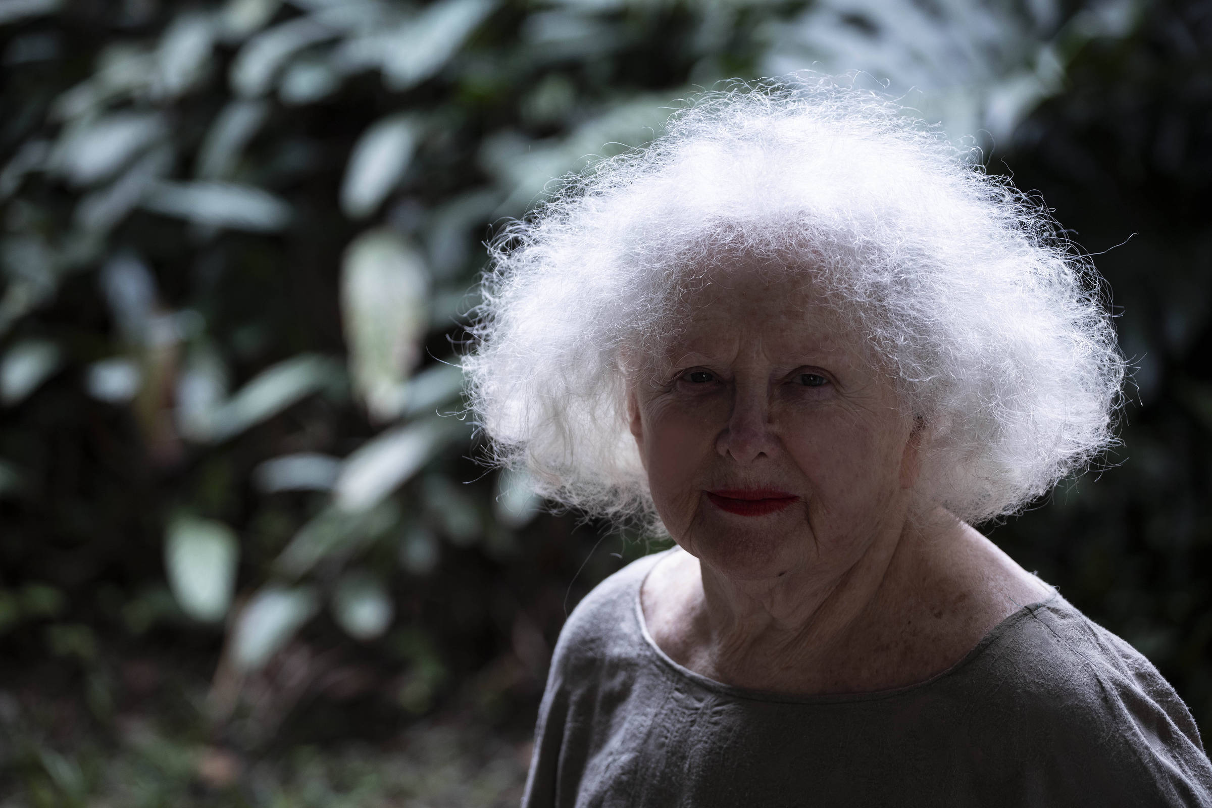 A imagem mostra uma mulher idosa com cabelo grisalho e volumoso, que está em um ambiente natural com vegetação ao fundo. Ela tem um olhar sereno e usa um vestido de cor neutra. A iluminação é suave, destacando seu rosto e cabelo.