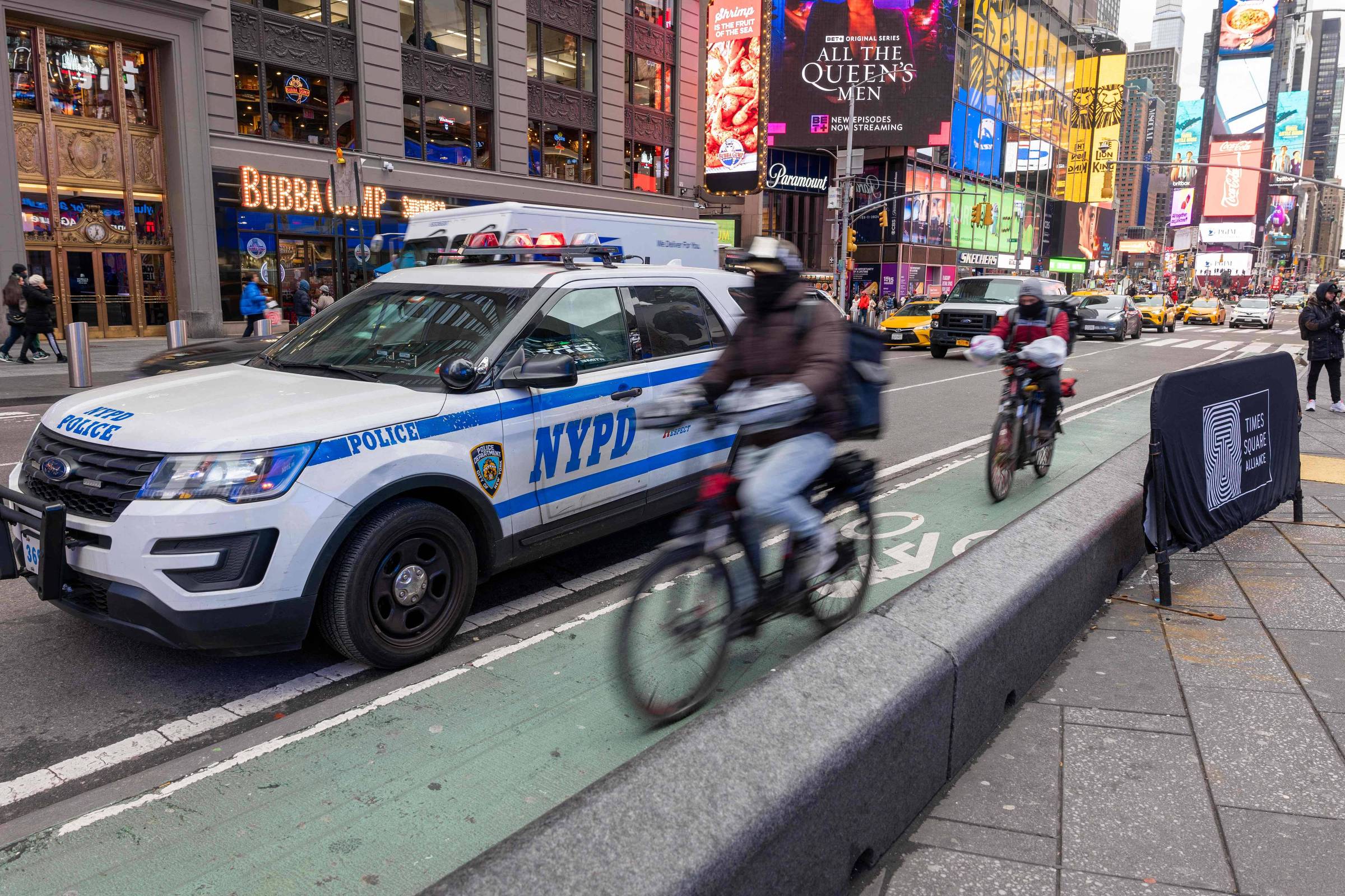 A imagem mostra uma cena em Times Square, Nova Iorque, com um veículo da polícia NYPD estacionado em uma faixa de ciclistas. Dois ciclistas estão passando ao lado do carro. Ao fundo, há letreiros luminosos e lojas, criando um ambiente urbano movimentado. A calçada é larga e há uma estrutura de proteção para ciclistas.