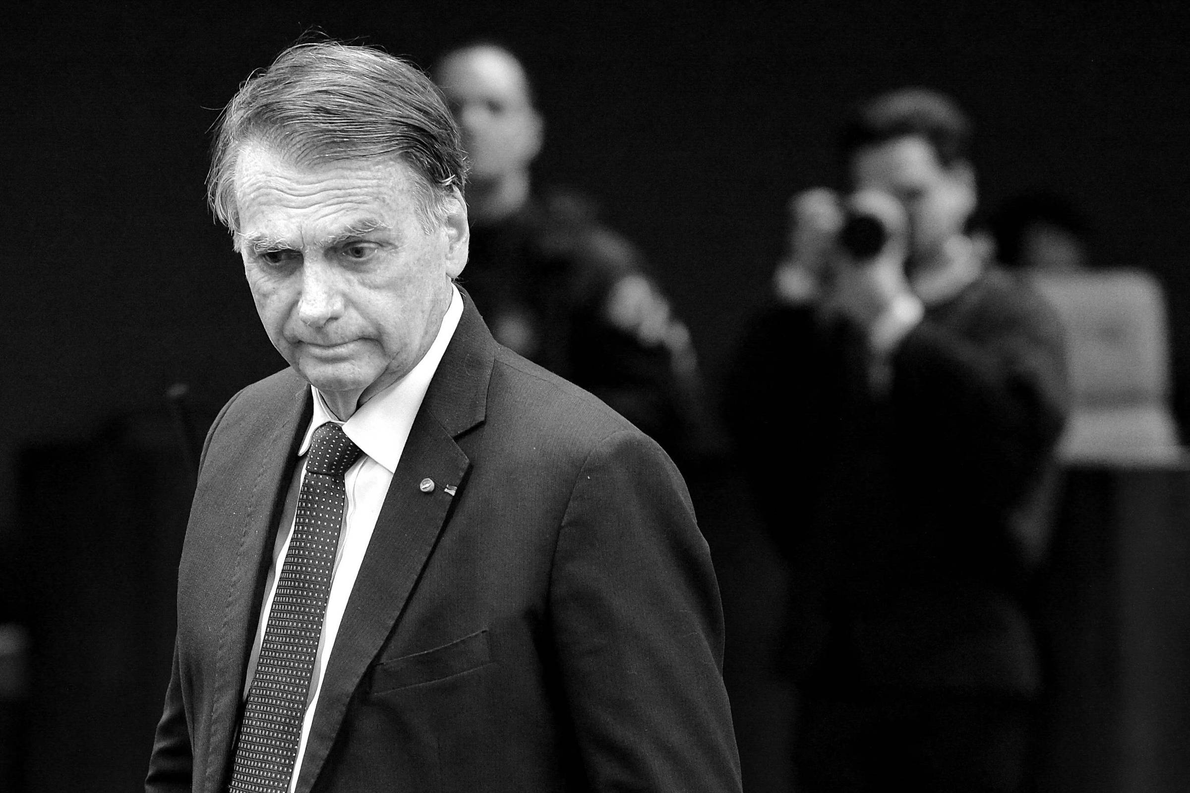 A imagem mostra um homem de terno escuro e gravata, com expressão séria, caminhando em um ambiente que parece ser uma sala de reuniões ou um plenário. Ao fundo, há pessoas, incluindo um homem com uma câmera, que parece estar tirando fotos. O homem em primeiro plano tem cabelo grisalho e está olhando para baixo, transmitindo uma sensação de introspecção ou preocupação.