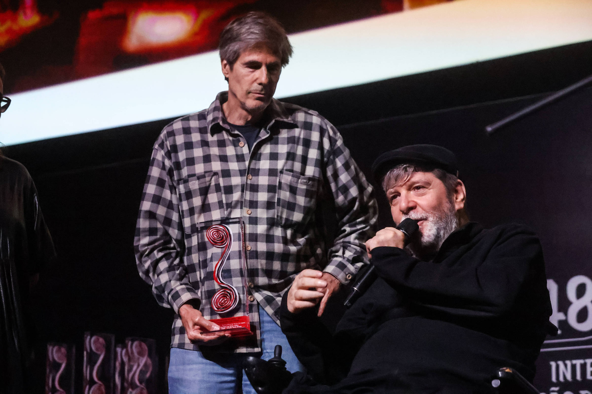 A imagem mostra duas pessoas em um palco durante uma cerimônia de premiação. À esquerda, uma pessoa está segurando um troféu, enquanto à direita, outra pessoa, que está em uma cadeira de rodas, fala ao microfone. O fundo é decorado com elementos gráficos relacionados ao evento.