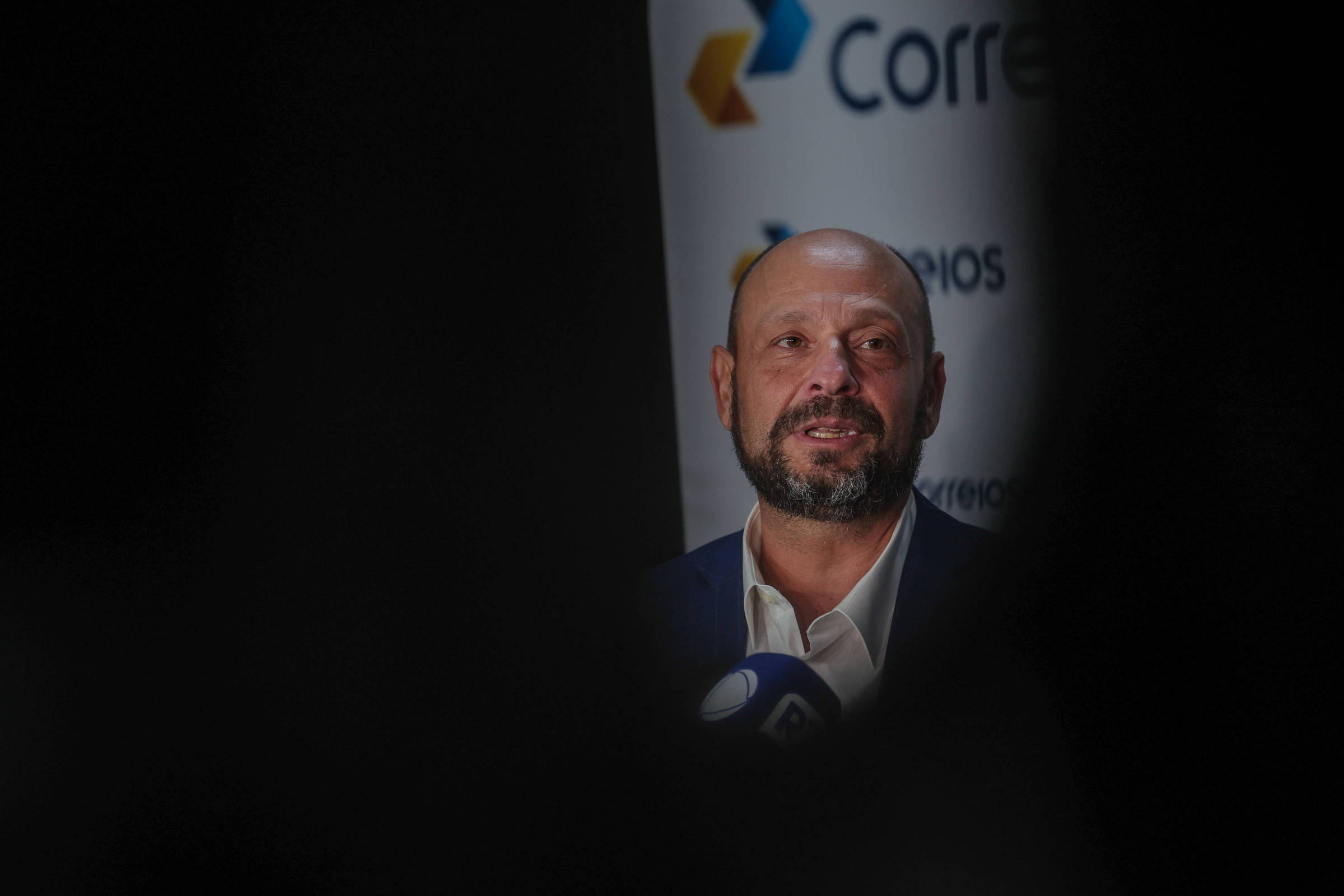 Um homem com cabelo curto e barba está falando em um evento. Ele usa um paletó escuro e uma camisa branca. Ao fundo, há um banner com o logotipo dos Correios e o texto