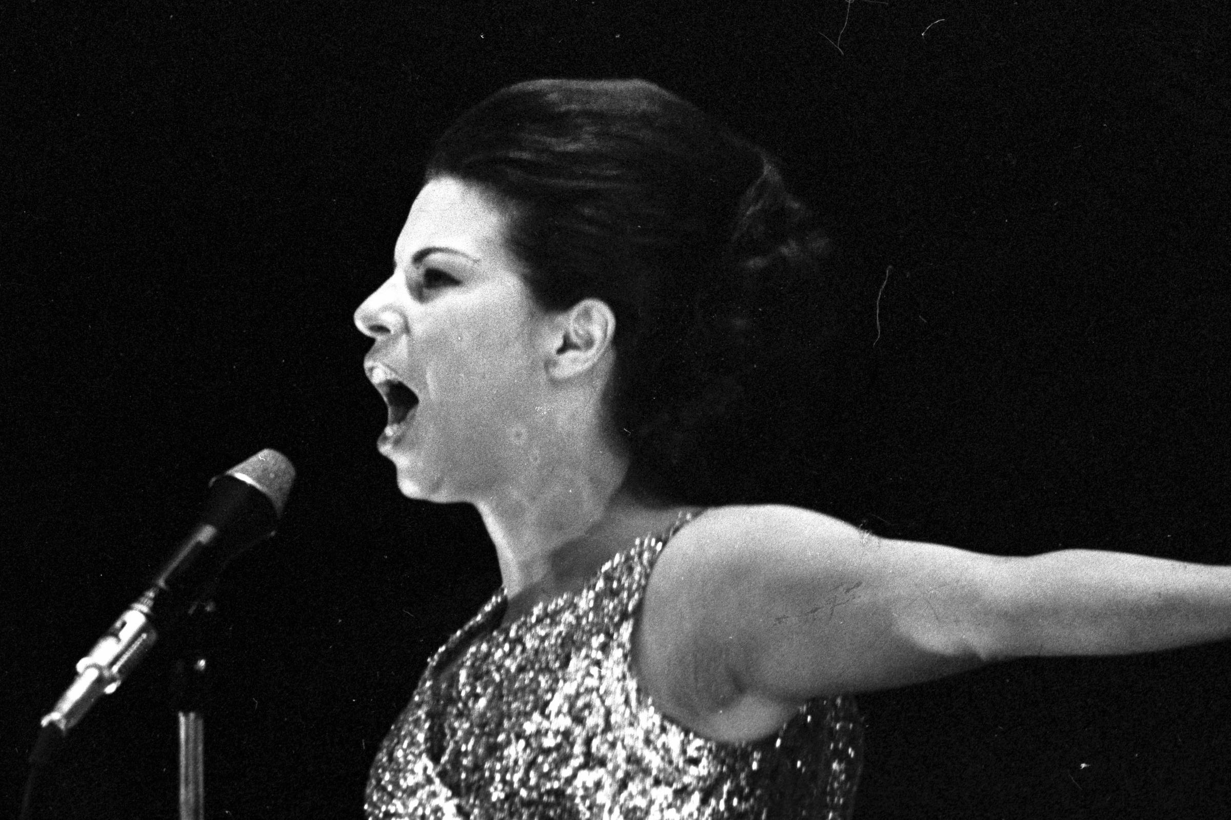 A imagem mostra uma mulher cantando em um microfone, com uma expressão intensa e emocional. Ela está vestida com um vestido brilhante e possui o cabelo preso em um coque. A iluminação é suave, destacando sua figura contra um fundo escuro.
