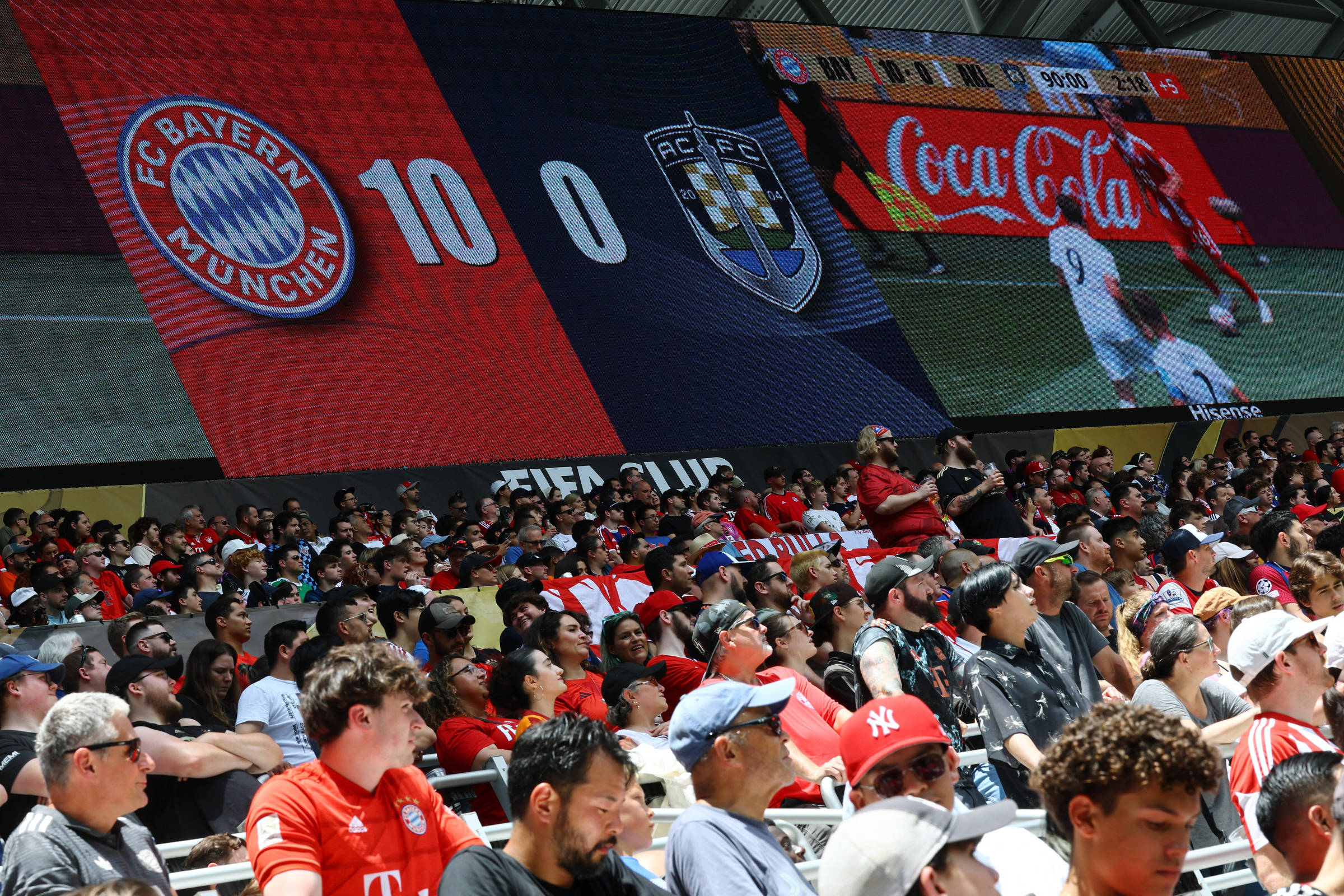 A imagem mostra uma grande tela de LED exibindo o placar de um jogo de futebol, onde o FC Bayern Munchen está vencendo por 10 a 0 contra o RC Porto. Abaixo da tela, há uma multidão de torcedores assistindo ao jogo, com muitos usando camisas vermelhas e brancas, típicas do Bayern. O ambiente é ensolarado e a atmosfera é de celebração.