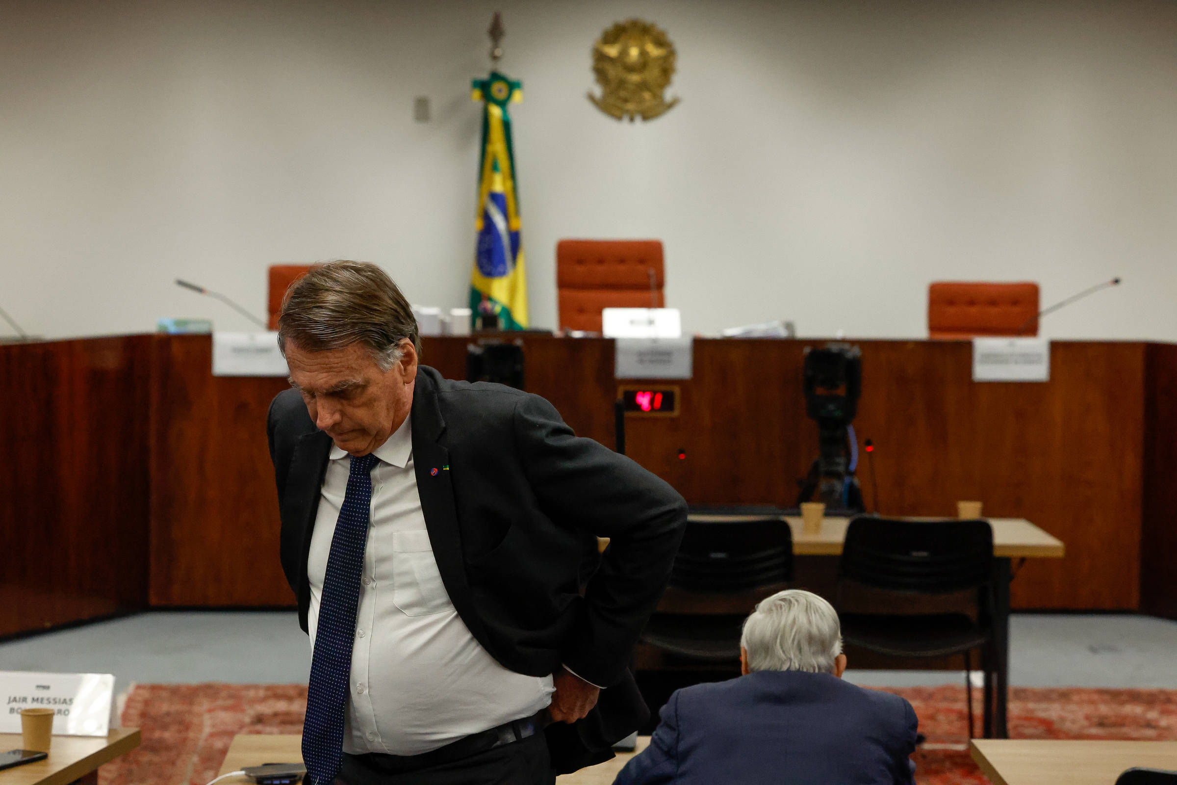 A imagem mostra um ambiente de tribunal com um homem de terno escuro, que parece estar se afastando, e outro homem sentado à mesa. Ao fundo, há uma bandeira do Brasil e o brasão nacional. As cadeiras do tribunal estão vazias, e há placas com nomes na mesa.