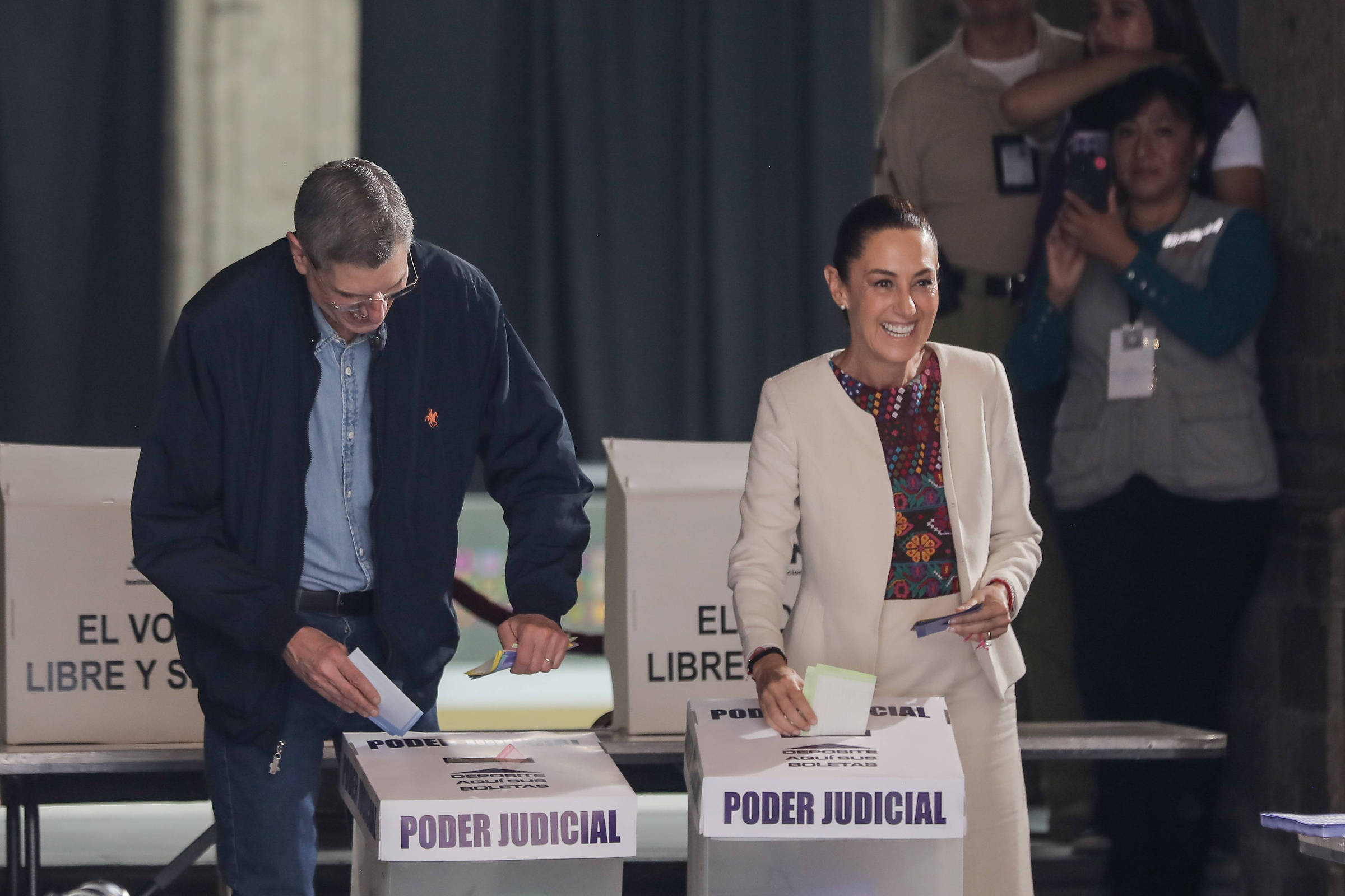 A imagem mostra duas pessoas participando de uma votação. À esquerda, um homem com cabelo curto e vestindo uma jaqueta azul escura e uma camiseta clara, está colocando um voto em uma urna. À direita, uma mulher com cabelo preso, vestindo um traje claro e uma blusa colorida, sorri enquanto segura um papel na mão. Ao fundo, outras pessoas observam a cena. As urnas têm a inscrição